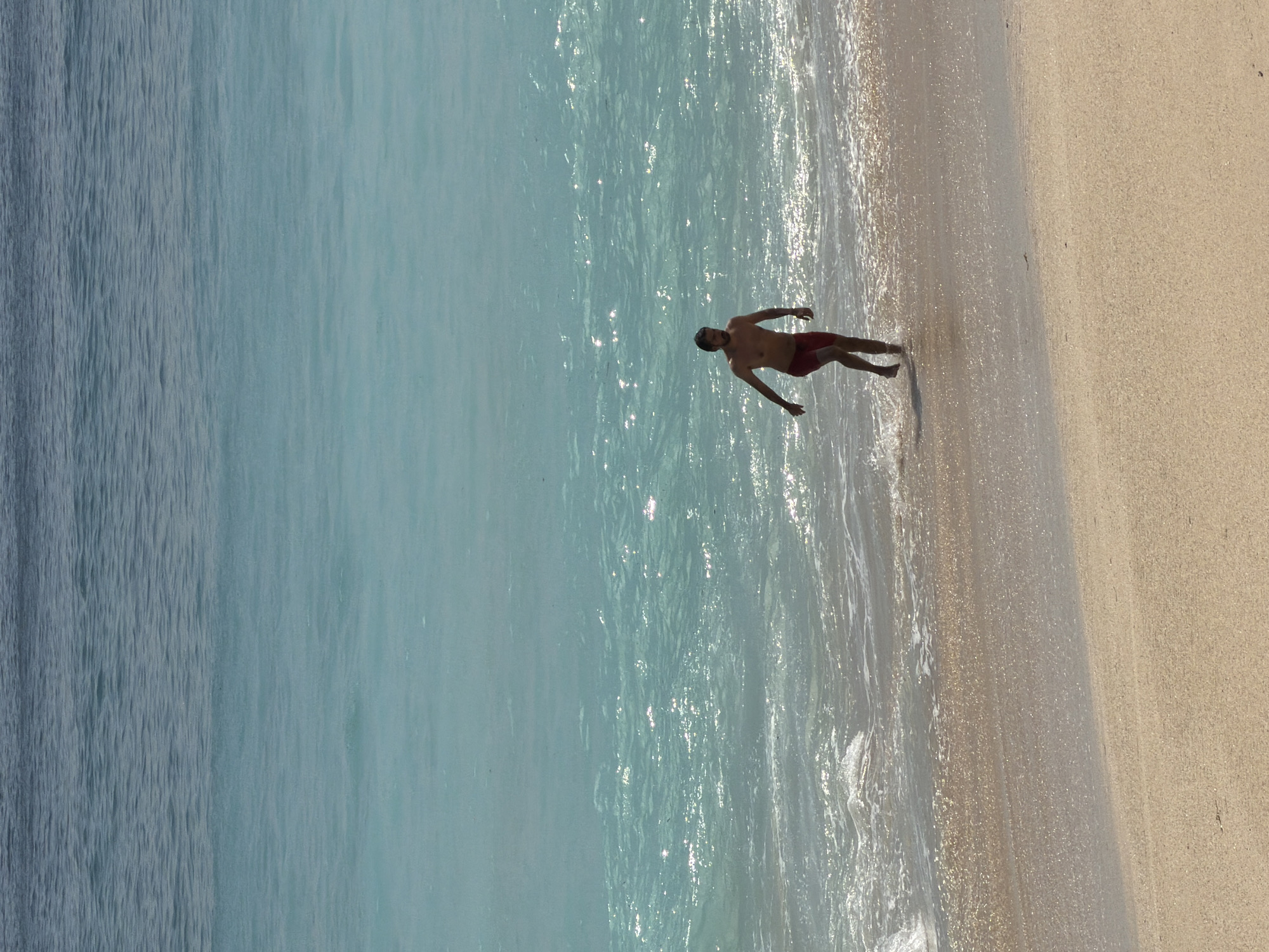 A man in red swim trunks walks through shallow waves on a sandy beach. The ocean is calm with gentle waves, and the water color transitions from deep blue in the distance to a bright turquoise closer to shore, reflecting sunlight. The sand is light brown and appears wet near the water's edge. The scene is an outdoor environment, likely a tropical beach, given the water colors and bright sunlight. The man is the sole subject in the frame, walking from right to left as the viewer perceives the image. The background is predominantly the ocean, with a darker blue band suggesting a deeper water area further out. There are no buildings or structures visible. The image appears to be a candid shot of a person enjoying the beach.