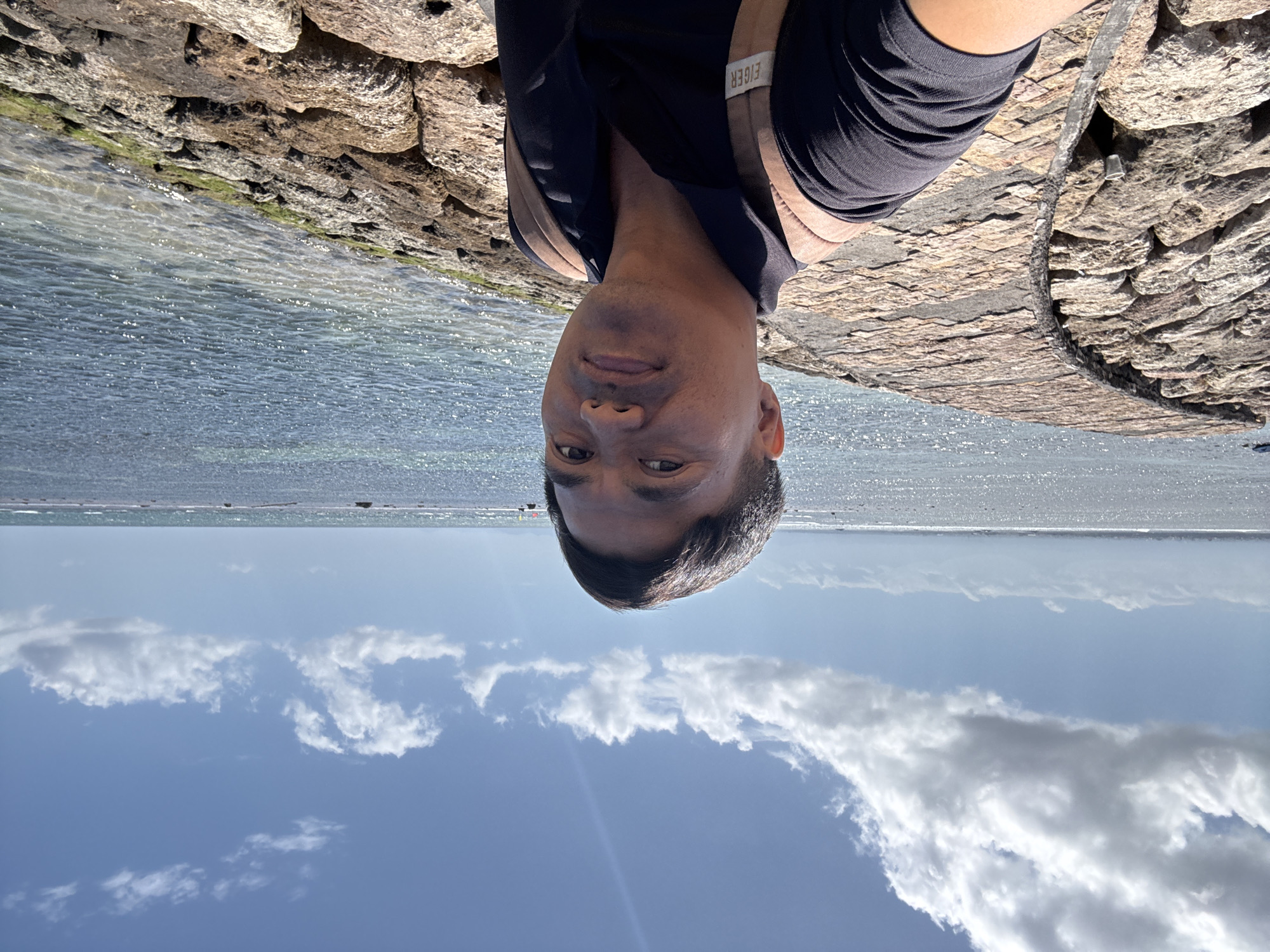 A man in a black collared shirt and a tan backpack takes a selfie on a rocky shore with a curved pathway made of stone slabs. The ocean is to his right, reflecting the blue sky and scattered white clouds. The sun is bright, casting highlights on the water. The pathway is constructed from rough-hewn, tan-colored rocks, and it curves away from the viewer to the left. Beyond the pathway, more rocks are visible, some covered in green algae. The man's face is clearly visible, and he appears to be looking directly at the camera. The background shows a clear horizon where the ocean meets the sky. The image appears to have been taken on a sunny day. This scene is consistent with the coastline in Denpasar, Indonesia, possibly near a breakwater or seawall.