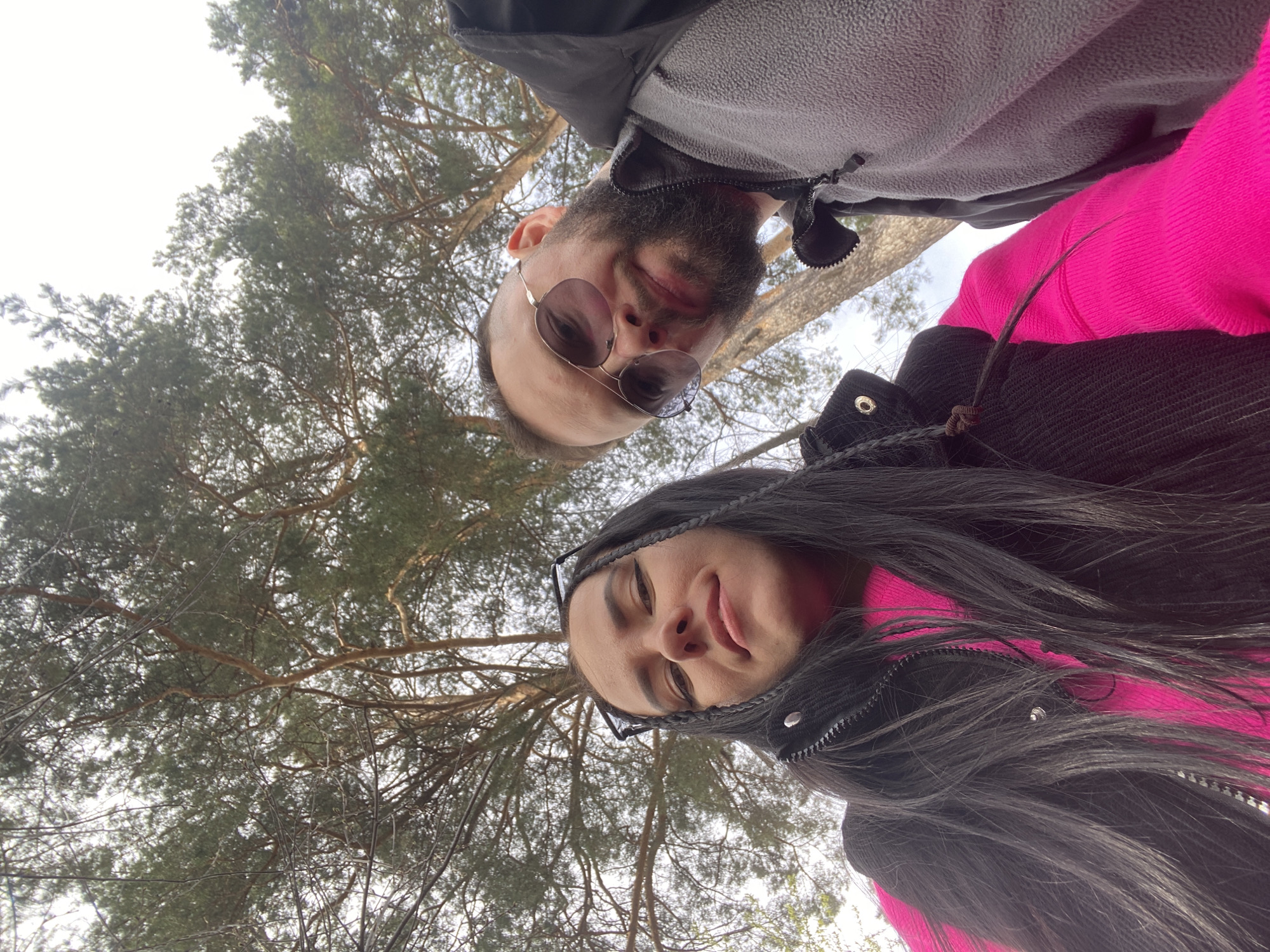 A low-angle shot depicts two individuals, a man and a woman, with a backdrop of a dense canopy of coniferous trees. The man, positioned slightly above and to the left, has short dark hair, a beard, and sunglasses. He is wearing a gray fleece jacket. The woman, below and to the right, has long dark hair, a pink sweater under a black jacket, and eyeglasses pushed atop her head. The trees form a dense overhead background with branches and needles filling most of the frame. Based on the landscape details, specifically the type of coniferous trees, and assuming the user's provided location data is correct, the photo was taken in Chaikovsky, Russia.