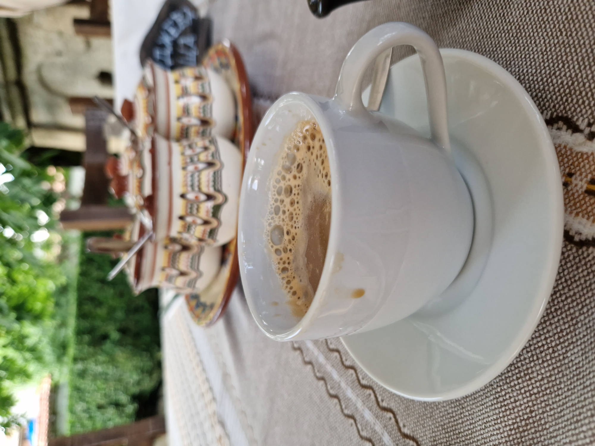 A close-up shot focuses on a white ceramic cup and saucer filled with a frothy beverage, likely coffee. The cup is positioned on the right side of the frame, with its handle clearly visible. Behind the cup, a decorative ceramic plate holds three lidded containers, possibly for sugar or jam, each adorned with colorful geometric patterns. These containers are also out of focus. The table is covered with a textured, neutral-colored tablecloth. In the background, a blurred landscape suggests an outdoor setting, with greenery and architectural elements, hinting at a restaurant or cafe in Panagyurishte, Bulgaria, possibly the "House of Radina" or a similar historical complex, known for traditional Bulgarian architecture and cuisine. The overall scene evokes a sense of leisure and enjoyment of a traditional beverage.