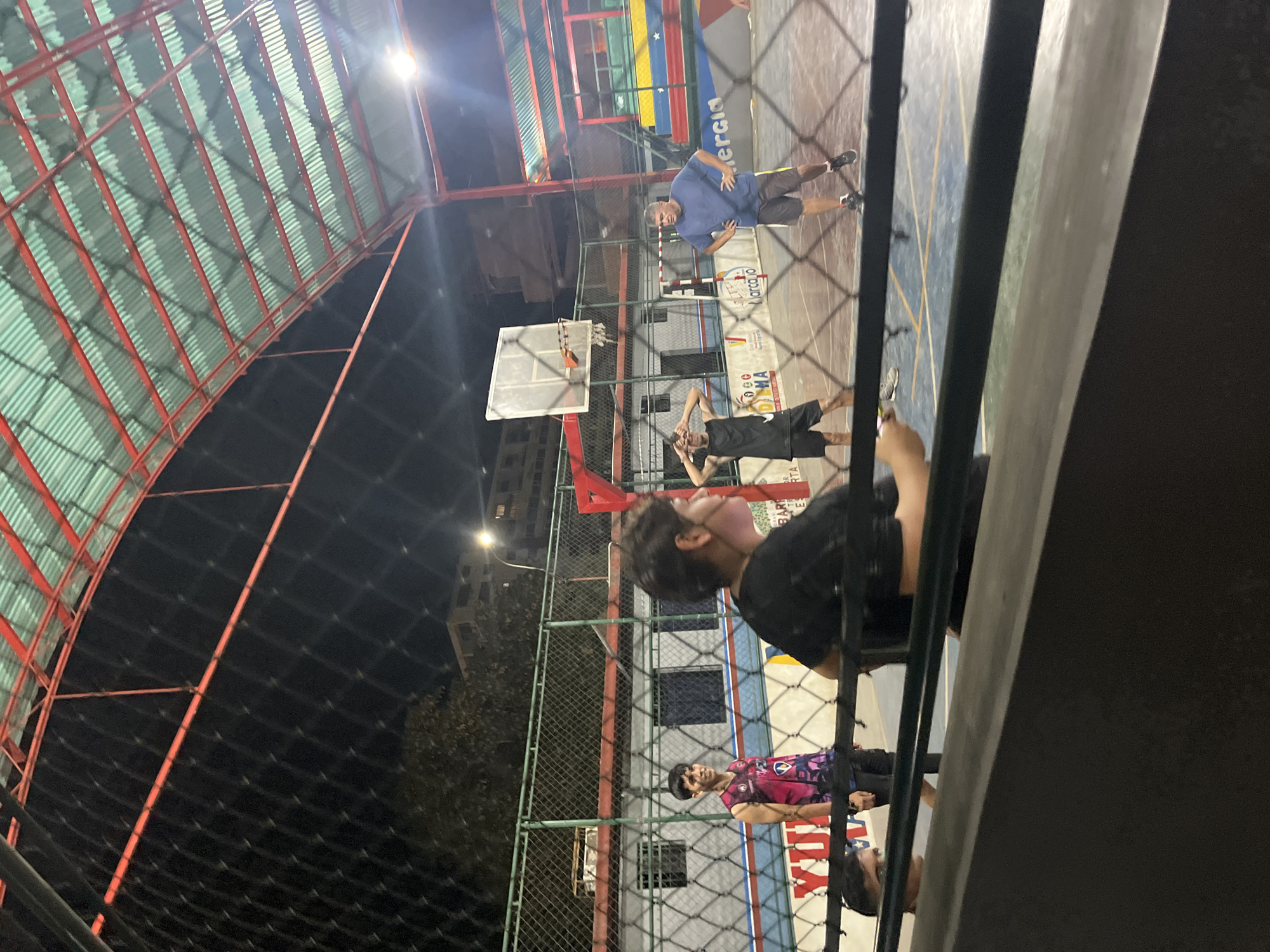 The image depicts an outdoor basketball court in Juan Griego, Venezuela. The court is enclosed by a chain-link fence and has a corrugated metal roof supported by red metal beams. Several people are present on the court: one man is shooting a basketball, another is observing, and two more are standing near the fence. The court features markings and the colors of the Venezuelan flag. The lighting suggests it is nighttime. Beyond the court enclosure, buildings and trees can be seen in the distance. The image is taken from behind a set of vertical bars, partially obstructing the view.