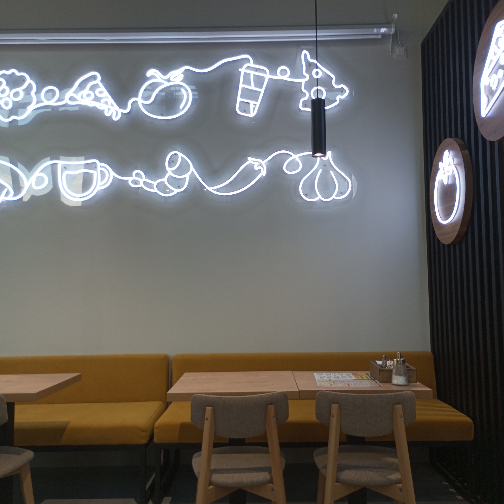 An interior shot captures a dining area with yellow booth seating and wooden chairs at a table. The wall behind the seating features a white neon sign depicting food items like pizza, a coffee cup, and vegetables, and a dark cylindrical pendant light hangs from the ceiling. To the right, a dark slatted wall has two round wooden signs with illuminated outlines of food, one resembling cheese and the other a tomato. The table has a menu and a salt and pepper shaker set. The scene is static and does not include any people.