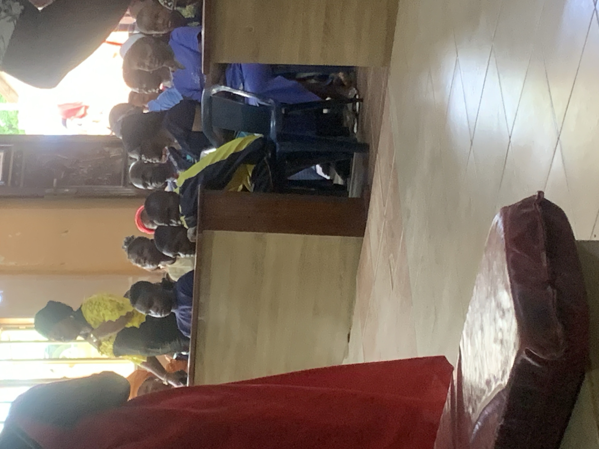 The image shows a group of people sitting in what appears to be a church or meeting hall. They are seated in rows, and the perspective is from the side, with a portion of a red and brown object in the foreground. People are dressed in various clothing, including blue, yellow, and black attire. There is a window or doorway visible in the background. The location is purportedly Akpaka Forest Reserve, Nigeria, although this cannot be confirmed visually from the image itself. The room has light-colored walls and tiled flooring.