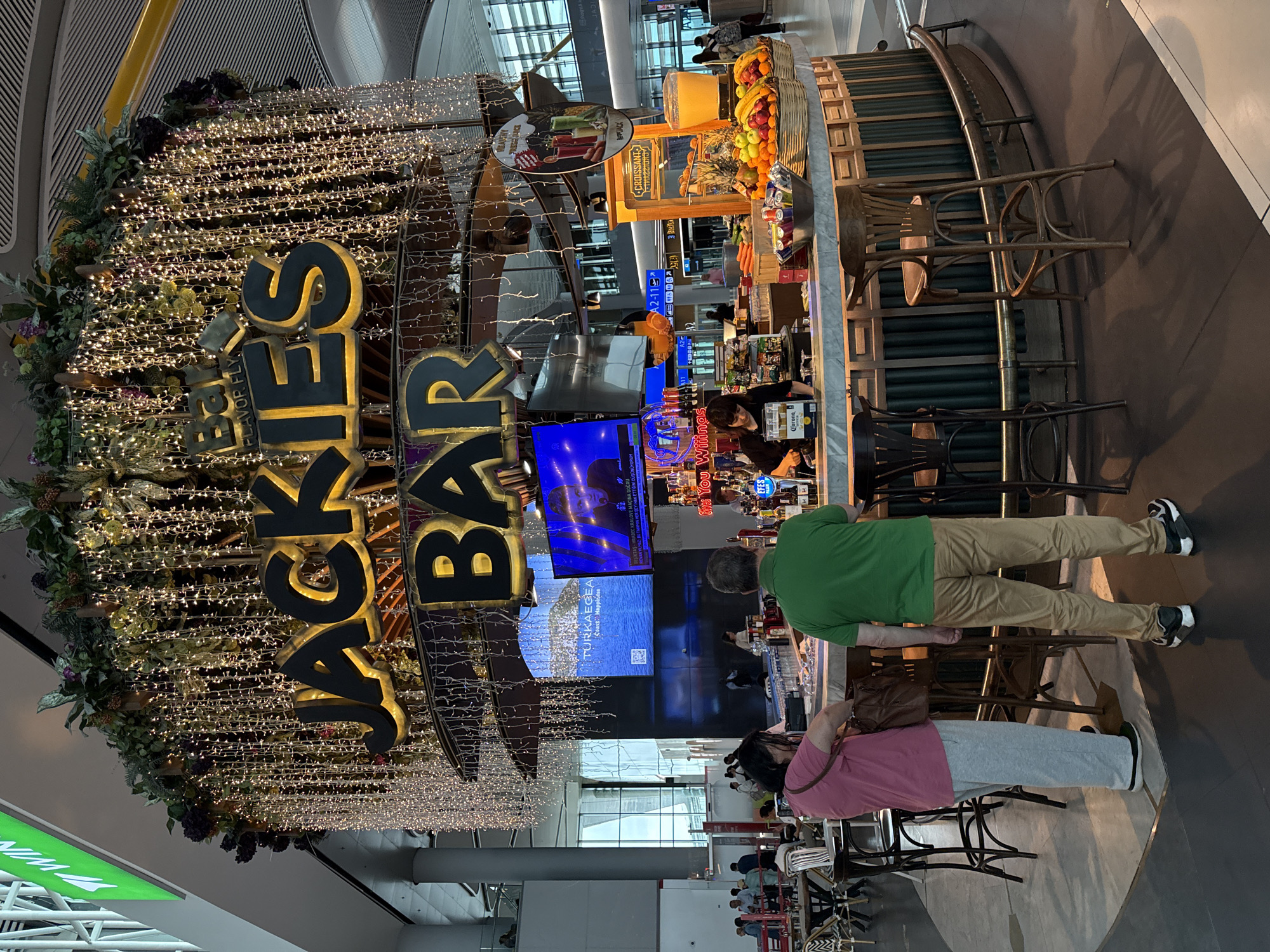 The image shows the interior of a bar named "Jackie's Bar" located in Arnavutköy, Turkey. The bar is decorated with numerous fairy lights and greenery, creating a festive atmosphere. A large sign with "JACKIE'S BAR" in bold yellow lettering is prominently displayed above the bar area.
In the foreground, two individuals, a woman in a pink shirt and grey sweatpants and a man in a green t-shirt and tan pants, are standing at the bar. The woman is looking down at her bag, while the man is facing the bar. A bartender, dressed in black, is visible behind the counter, attending to drinks.
The bar itself is circular and features a marble countertop. Behind the counter, there are shelves stocked with bottles of alcohol, a beer dispenser, and other bar essentials. To the right of the bar, there's a display of fresh fruits and juices.
In the background, a large screen displays images of a coastal landscape and news content. The overall environment appears to be an indoor public space, possibly an airport terminal or a large entertainment venue, given the modern architecture and the presence of other patrons and seating areas visible in the distance. The lighting suggests it is daytime, but the decorative lights of the bar are also illuminated.