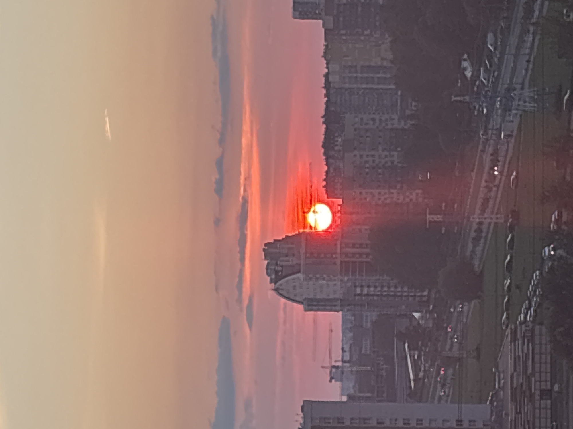 The image depicts a sunset over a city, likely in Russia based on the context. The sun, a bright orange-red orb, is partially obscured by clouds and is positioned centrally in the frame, just above a cluster of buildings. The sky is a gradient of warm colors, transitioning from a soft orange at the top to a vibrant pink and red closer to the horizon.
In the foreground, a cityscape is visible. Tall residential buildings, some with distinctive architectural features like curved roofs, dominate the scene. The buildings appear to be modern, with visible windows and balconies. Below the buildings, a road is visible with a steady stream of car headlights and taillights indicating traffic flow. Trees and green spaces are interspersed between the buildings and the road, suggesting a urban environment with some landscaping. In the far background, several construction cranes are visible, hinting at ongoing development in the city. The overall impression is of an urban setting at dusk, bathed in the warm glow of the setting sun.