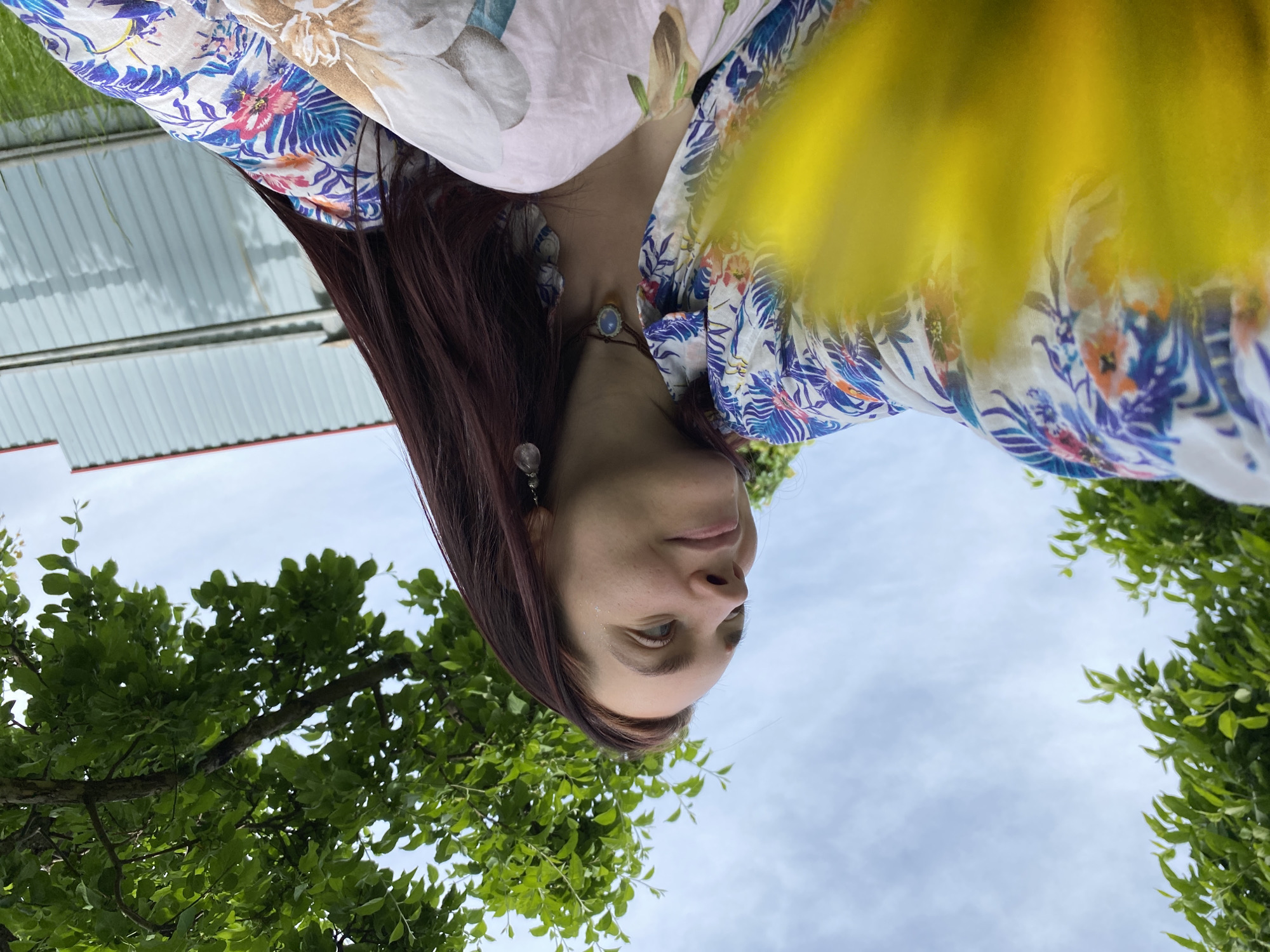 An outdoor portrait captures a young adult woman looking upwards, from a low-angle perspective. She is positioned on the right side of the frame, with her head slightly tilted. Her long, dark reddish-brown hair cascades over her shoulder. She is dressed in a light-colored top featuring a vivid floral pattern of blue, pink, and orange flowers and green leaves on a white background. Over this, a softer, flowing fabric with larger, muted floral designs in brown and light pink drapes across her. She wears a delicate necklace with a light blue or moonstone-like pendant and a silver-toned earring. Her expression appears calm and contemplative.
The background is dominated by a bright, partly cloudy sky. Lush green tree foliage, with visible leaves and branches, frames the lower left and parts of the upper left and lower right sections of the image. In the upper left distance, a light-colored building or fence with horizontal paneling is visible. A prominent, soft, bright yellow blur occupies the upper right portion of the image, possibly a close-up flower or a strong lens flare, obscuring part of her clothing and the sky. The natural lighting suggests it is daytime. No text is visible. The scene is set in an outdoor environment in Chaikovsky, Russia.