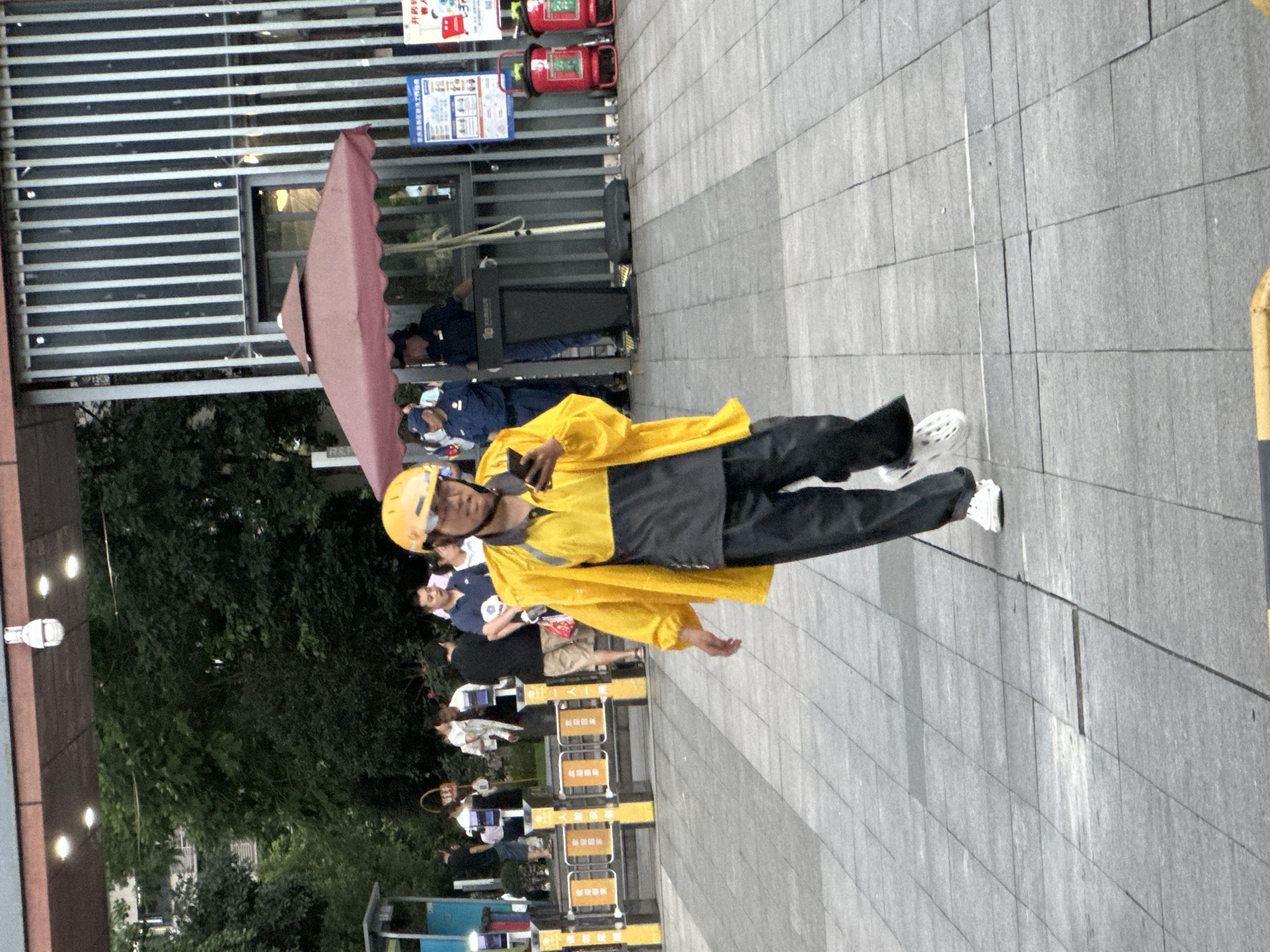 A person wearing a yellow jacket and helmet, possibly a delivery worker, is walking on a paved area in front of a building in Chongqing High-tech Zone, China. The person is holding a smartphone and looking at it. In the background, there are other people, a red umbrella, a security booth, and some signage. The environment appears to be an urban public space.