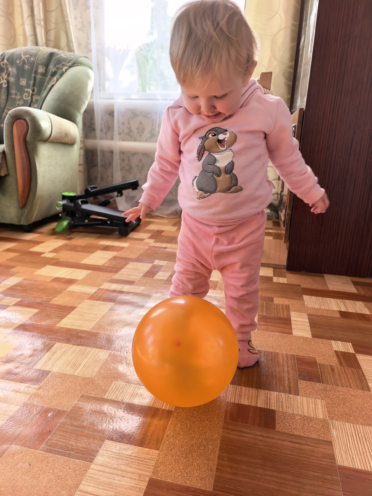 A young child, wearing a pink hoodie with a rabbit graphic and pink pants, stands on a patterned floor in what appears to be a living room. The child is looking down at a bright orange balloon in front of them. In the background, a green armchair with a patterned blanket is visible, along with a window with sheer curtains and a dark wooden cabinet. A piece of exercise equipment is situated behind the child. The floor has a wood-grain and cork tile pattern. There is no specific indication of location within Sterlitamak, Russia.