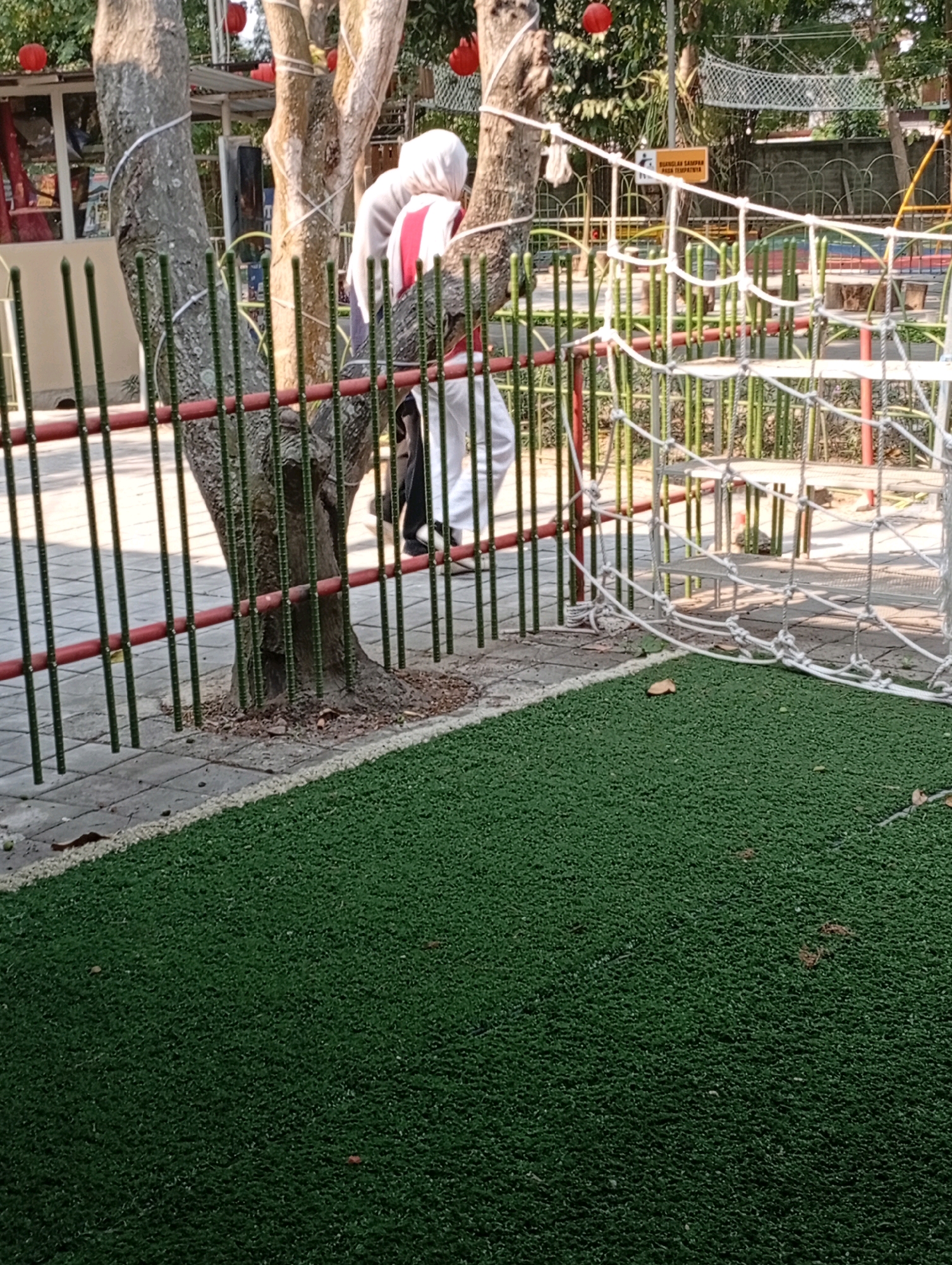 The image depicts an outdoor scene with artificial turf in the foreground and a children's playground in the background. In the mid-ground, a green metal fence with red accents separates the playground from the foreground. Two trees with white ropes wrapped around them stand behind the fence. Partially visible behind the trees are two individuals dressed in white and red. One individual wears a white head covering. The playground features a rope net structure and colorful playground equipment. A yellow sign with Indonesian text is visible on a pole in the background. The overall scene suggests a public park or recreational area. The location is identified as Pandaan, Indonesia.