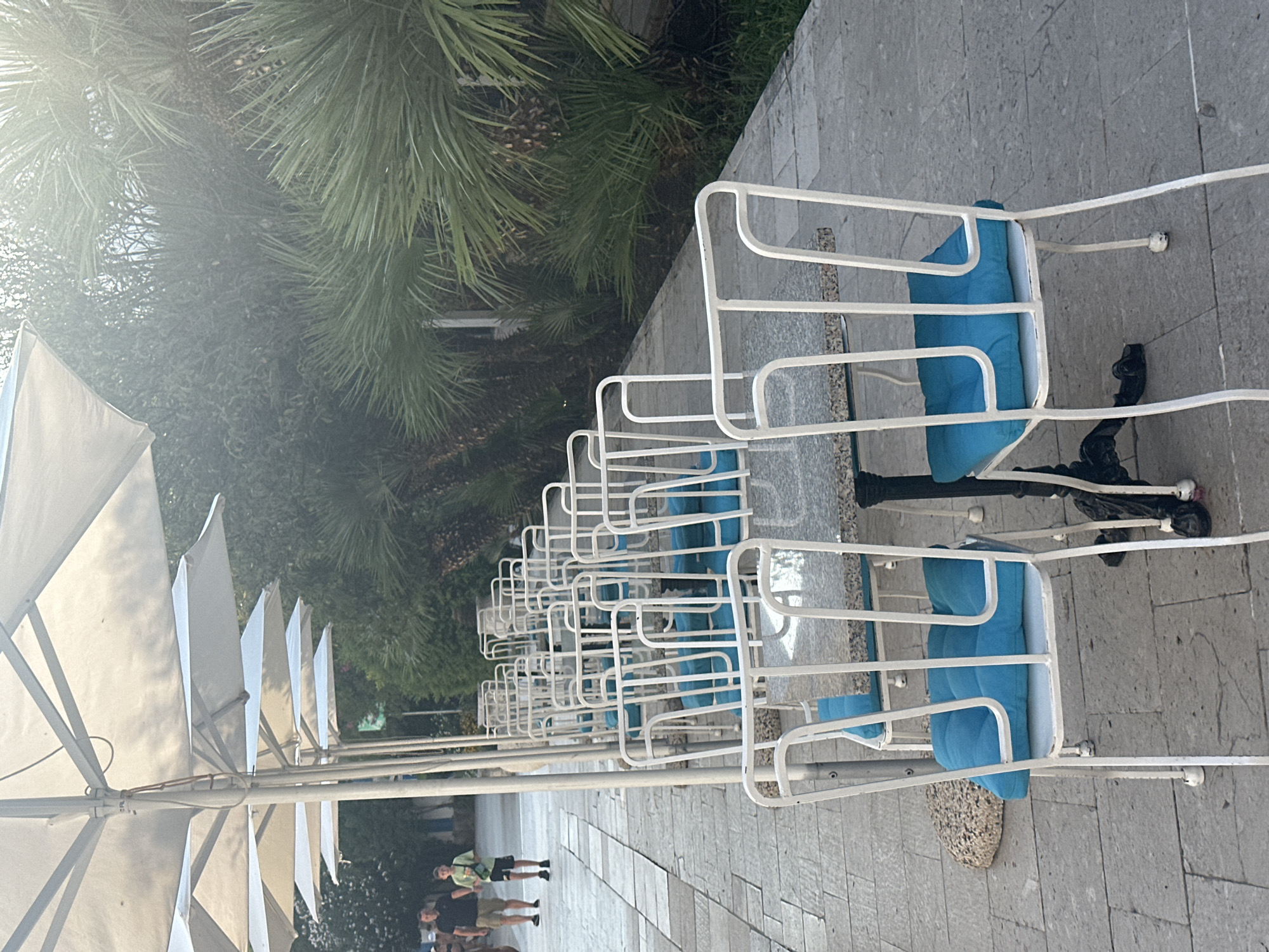 In Budva, Montenegro, a row of outdoor cafe tables with white chairs, each adorned with a bright blue cushion, are set up along a paved walkway. The tables have speckled granite tops and ornate black metal bases. Behind the tables, large white umbrellas are erected, providing shade. Beyond the umbrellas, lush green palm trees and other foliage create a verdant backdrop. In the distance, on the walkway, two individuals, likely tourists, are walking. The scene suggests a relaxed outdoor dining or seating area, possibly part of a restaurant or cafe. The sunlight filters through the trees, creating a slightly hazy effect in parts of the image.