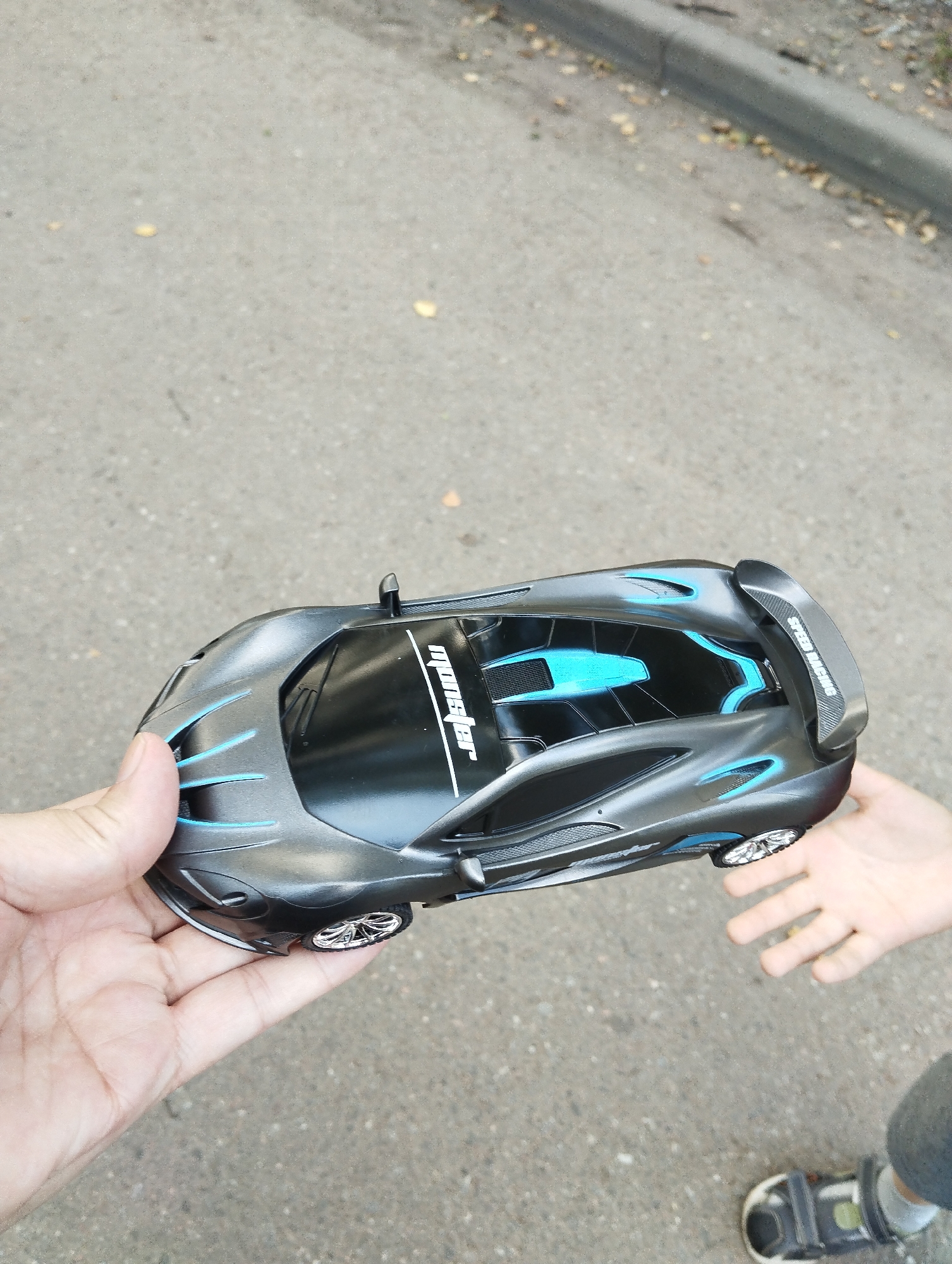 An adult's left hand holds a gray toy remote control car with blue accents and the text "SPEED RACING" on its rear spoiler. The car is positioned over a paved surface, possibly a sidewalk or road, with fallen leaves scattered around. To the right, a child's left hand is extended towards the car. In the lower right corner, a child's shoe with Velcro straps is visible. The background shows a gray curb and a sidewalk or road. The location is identified as Kingisepp, Russia.