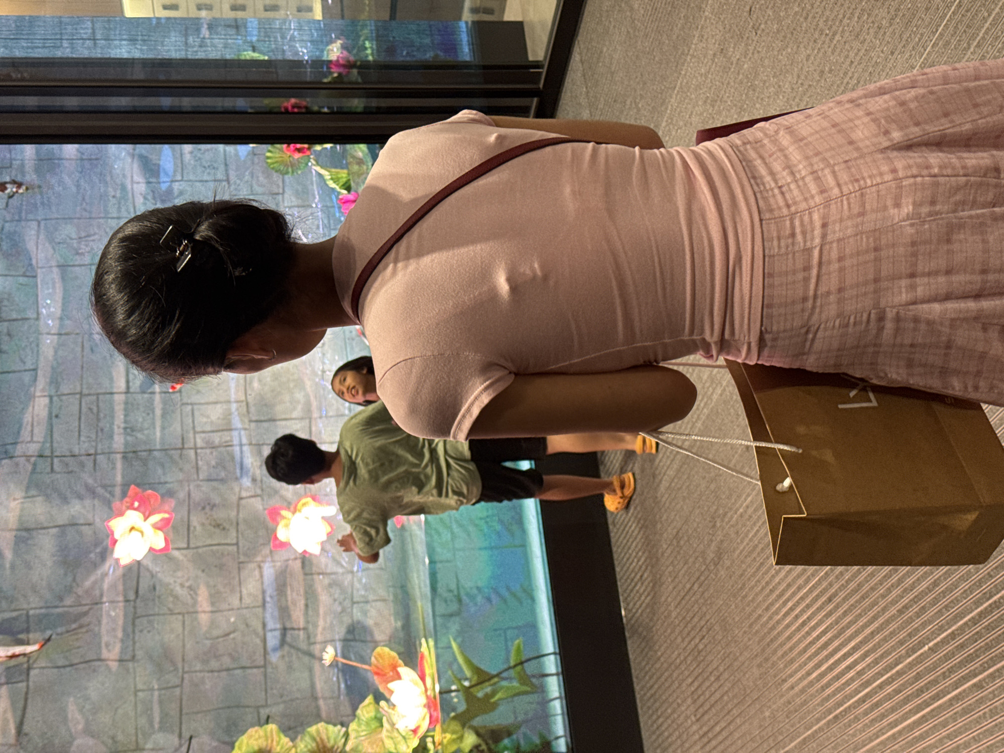 In a shopping mall in Hanoi, Vietnam, a woman in a pink outfit carries a shopping bag. In front of her, a boy in a green shirt and black shorts stands by an illuminated display of koi fish and lotus flowers, while a younger girl looks on. The display appears to be a large aquarium or a decorative wall feature. The flooring is carpeted with a striped pattern.