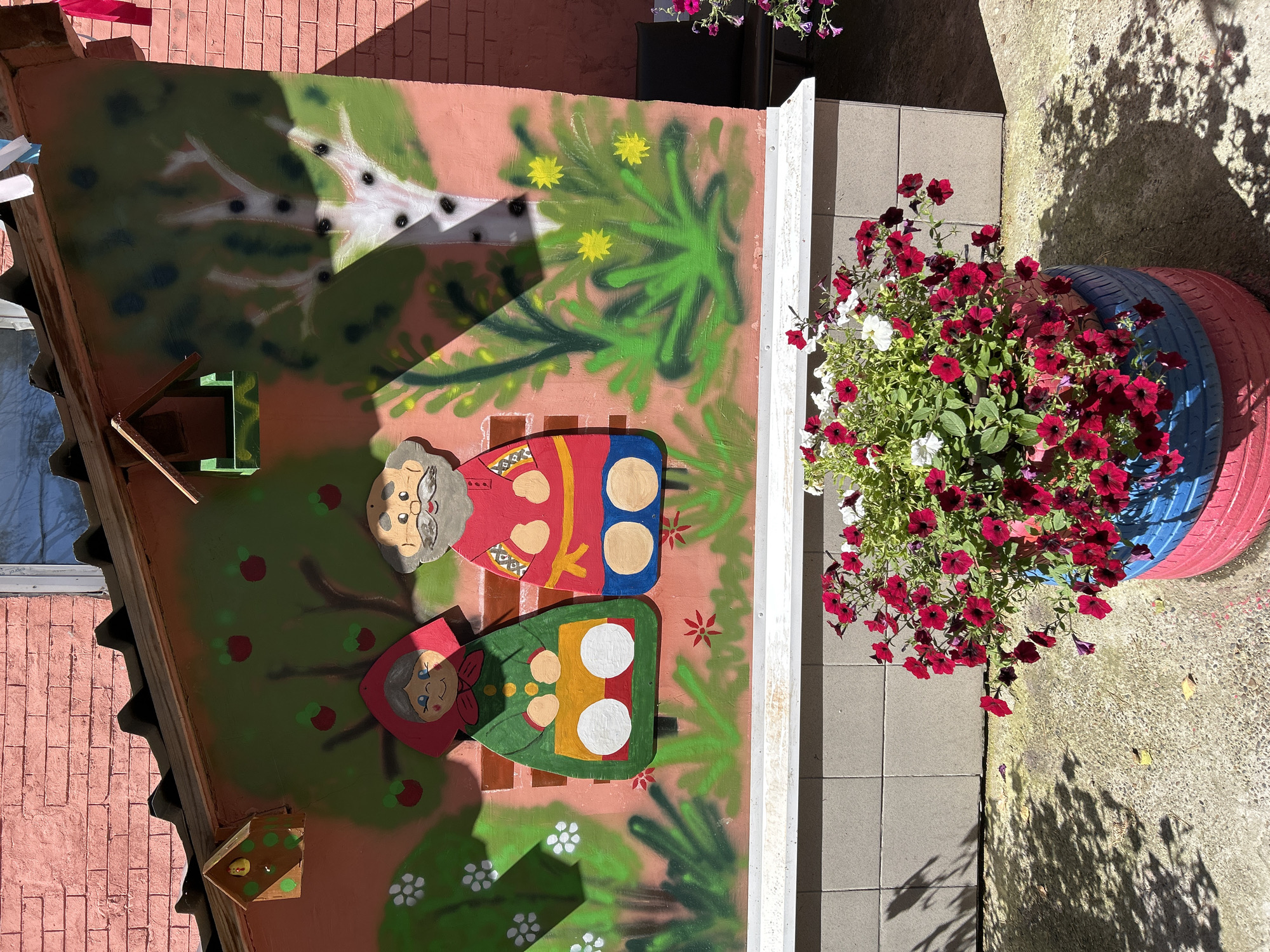 The image depicts a colorful outdoor mural painted on a pink brick wall, possibly part of a playground or community area. The mural features stylized representations of an elderly couple sitting on a bench, set against a backdrop of trees and flowers. To the left, a woman wears a red headscarf and a green dress with white circles. To the right, a man has a grey beard and wears a red tunic with decorative elements and blue trousers. A small birdhouse is attached to the roof overhang above the mural. In the foreground, a large planter, made from stacked and painted tires (red, blue, and pink), overflows with vibrant red and white petunias. The ground is paved with a light-colored, textured surface, with shadows of tree branches cast upon it, suggesting a sunny day. The overall scene is cheerful and decorative, likely intended to brighten a public space. The context of Tomsk, Russia, suggests a location in Siberia.