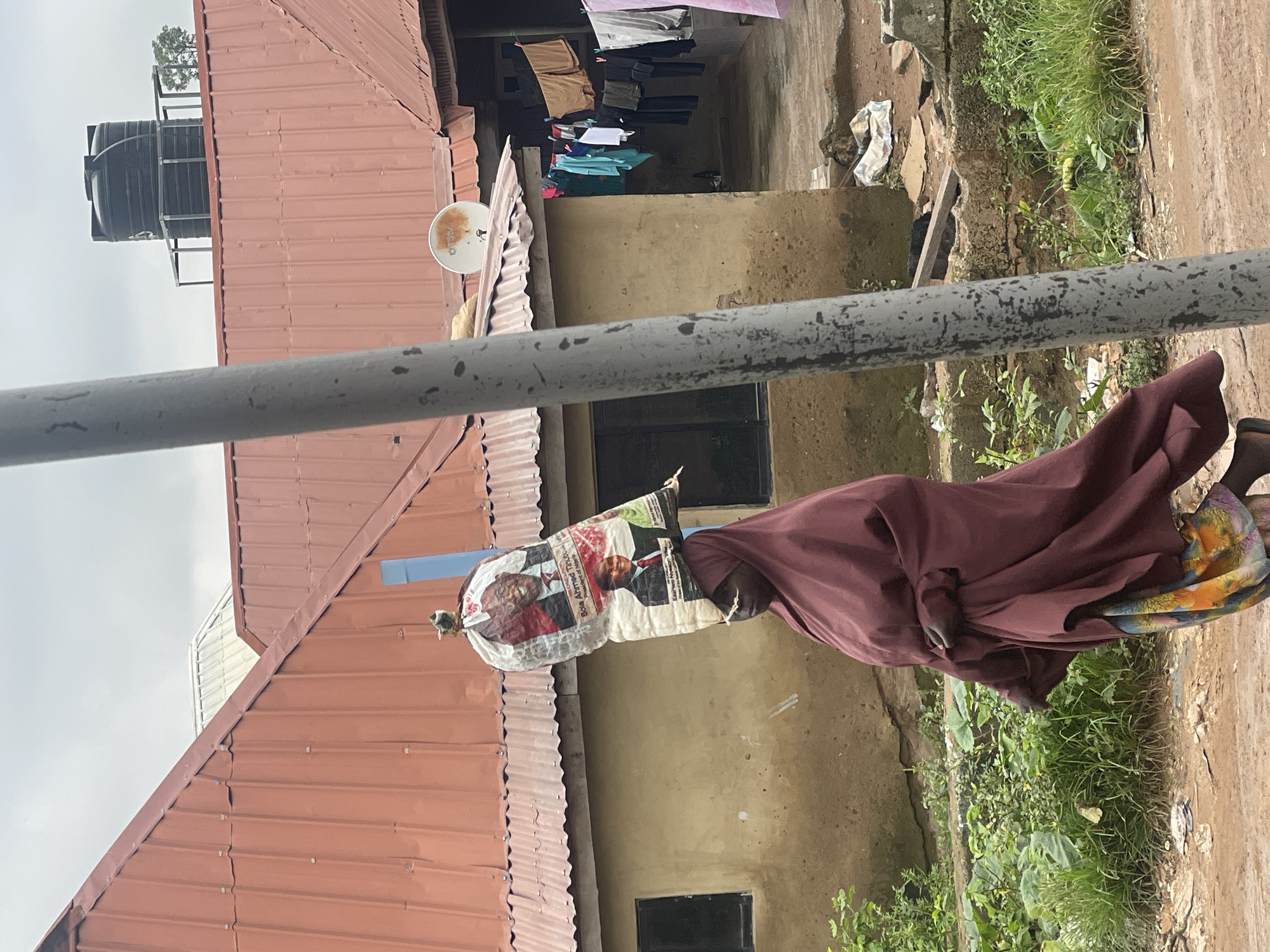 In the city of Kurunduma, Nigeria, a person is walking with a large bag balanced on their head. The bag is adorned with images of two men in suits, with text that appears to be political in nature. The person is wearing a burgundy headscarf and covering, and a brightly colored skirt is visible at the hem of their attire. They are walking on a dirt path, with overgrown green vegetation to the left and a building with a red corrugated metal roof to the right. The building has windows and laundry is hanging on a line outside. A satellite dish is mounted on the roof. A tall grey pole stands in the foreground, partially obscuring the view of the person. The sky is overcast.