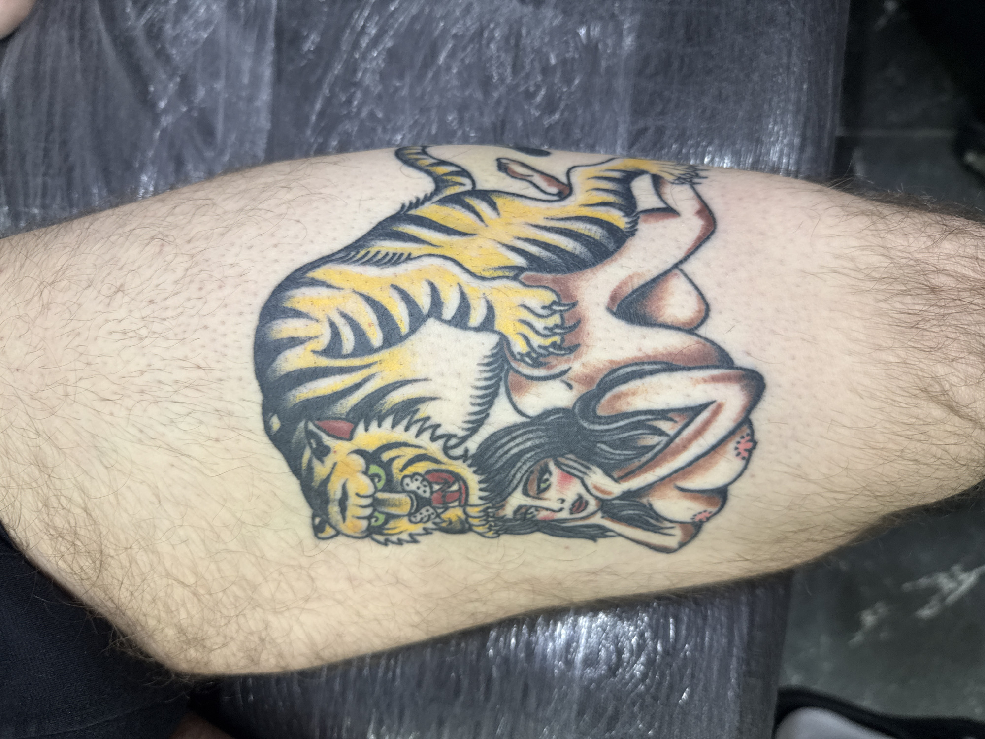 The image shows a tattoo on a person's hairy leg. The tattoo depicts a tiger entwined with a nude woman. The tiger is yellow with black stripes and green eyes, appearing to be snarling. The woman has black hair and is positioned beneath the tiger. The background is a mix of gray and black surfaces. There is no indication that the image was taken in Moscow, Russia.