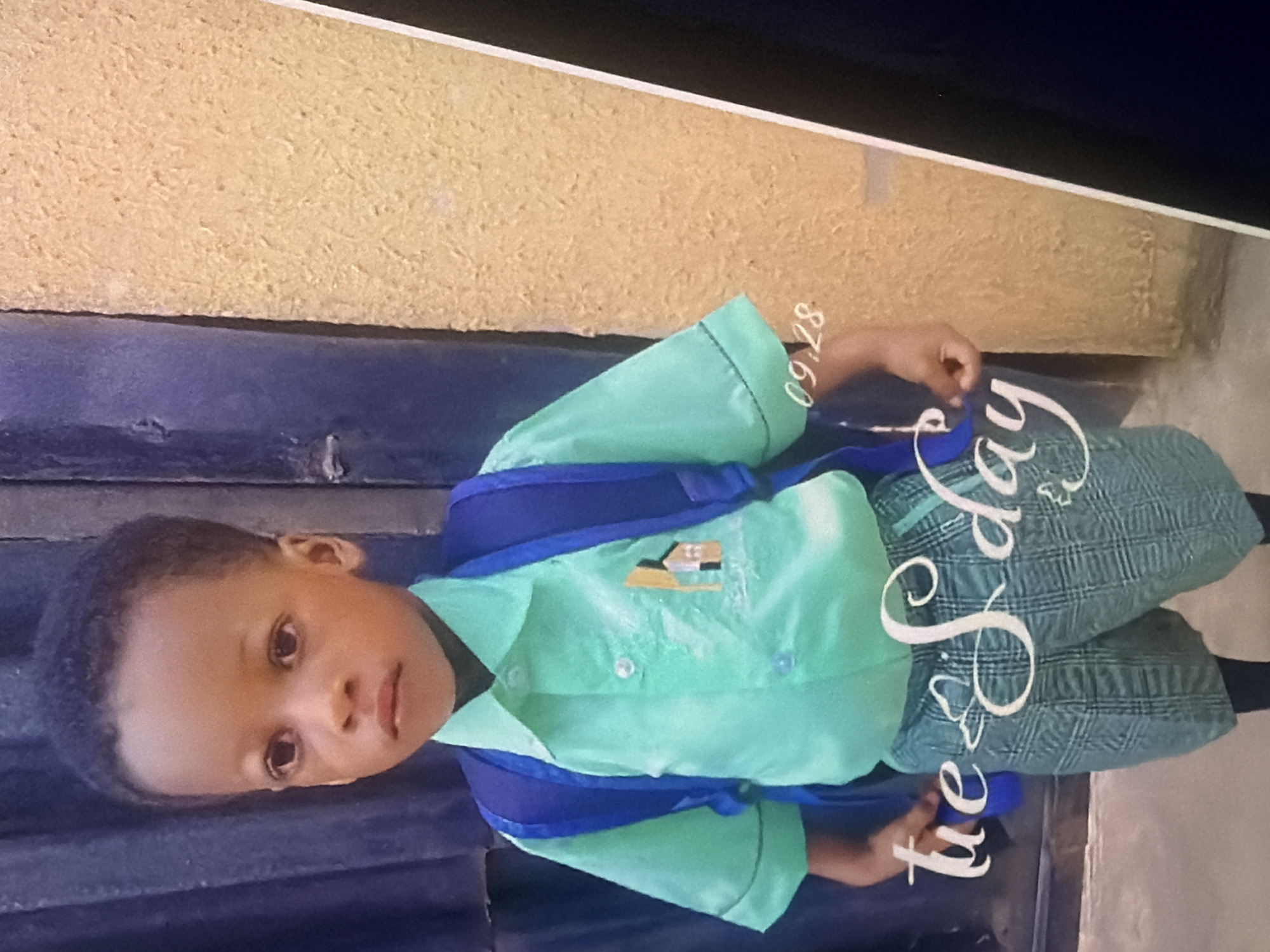 A young Black child, appearing to be a boy, stands facing forward in an outdoor setting, possibly a school entrance. He is wearing a light green button-down shirt with a small embroidered house graphic on the left breast, and plaid shorts over black leggings. A blue backpack is slung over his shoulders. The background consists of a dark blue corrugated metal door on the left and a textured yellow wall on the right. The image is overlaid with text that reads "tue s day" in a decorative white script, and the numbers "09.28" in a smaller font on the lower right. The child's expression is neutral, and his hands are by his sides.