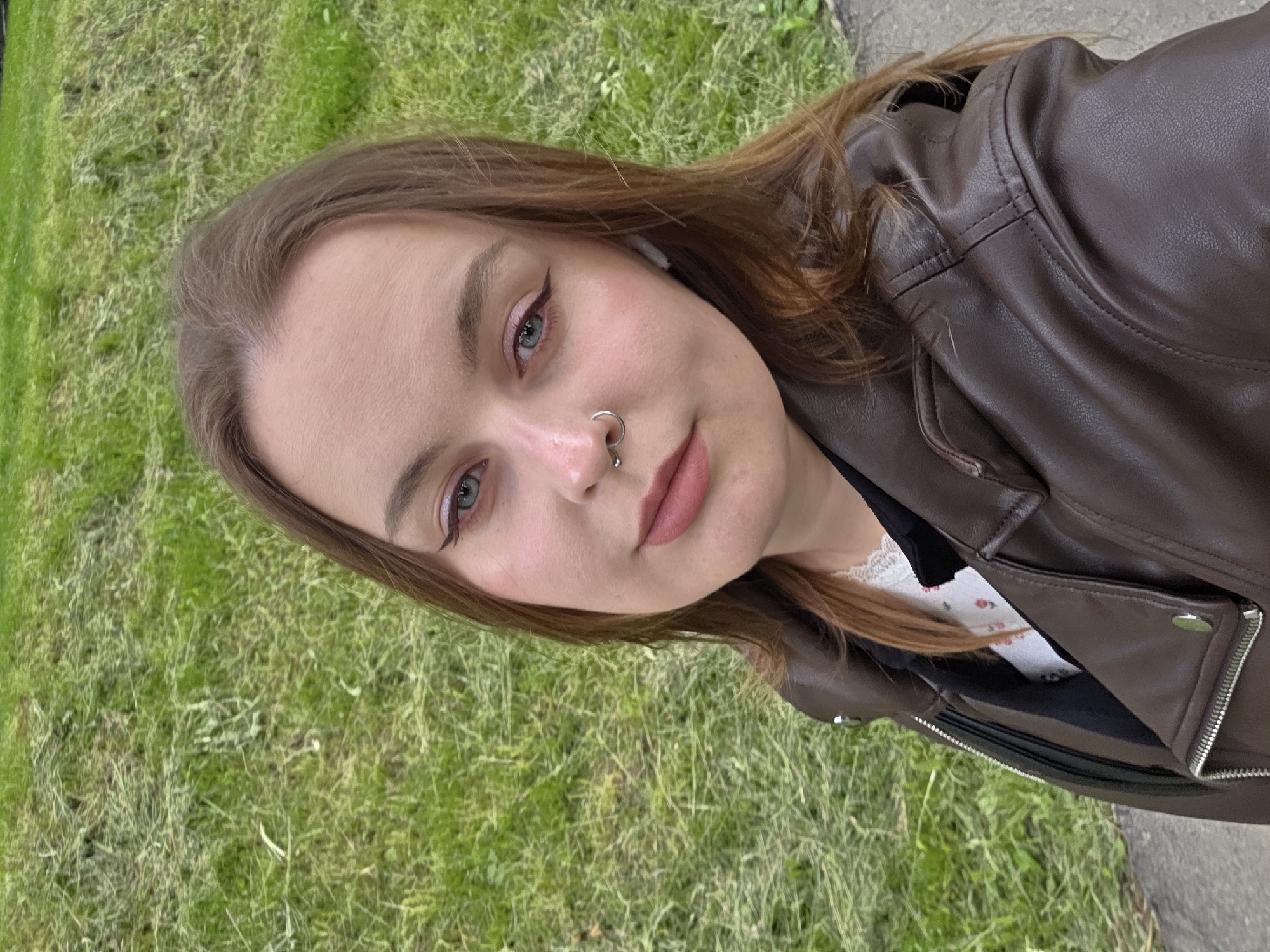 A young woman is taking a selfie outdoors. She is wearing a brown leather jacket over a white top with a floral print and a black underlayer. She has light brown hair, fair skin, and is wearing makeup, including eyeliner. She has a nose ring and an earbud in her ear. The background shows green grass and a grey pavement edge. The location appears to be an urban park setting.
