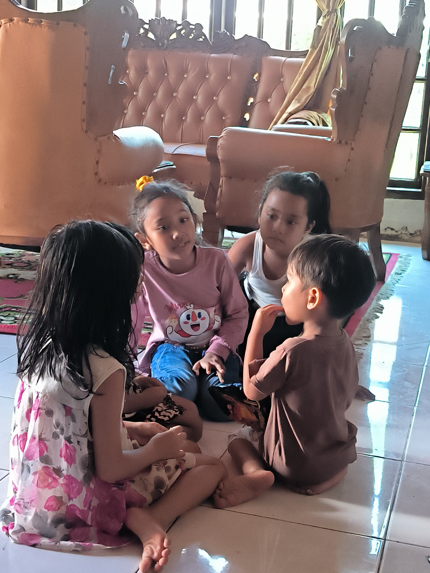 Four children are seated on a tiled floor in Merbopuntong, Indonesia. A girl in a floral dress faces away from the camera. A girl wearing a pink shirt and blue jeans faces her. Behind them is a girl wearing a white tank top. A boy in a brown shirt faces right, his hand raised to his mouth. The children sit in a circle. Behind the children is a brown leather sofa and a window with light curtains. A rug is beneath the sofa. The floor is light-colored and shiny.