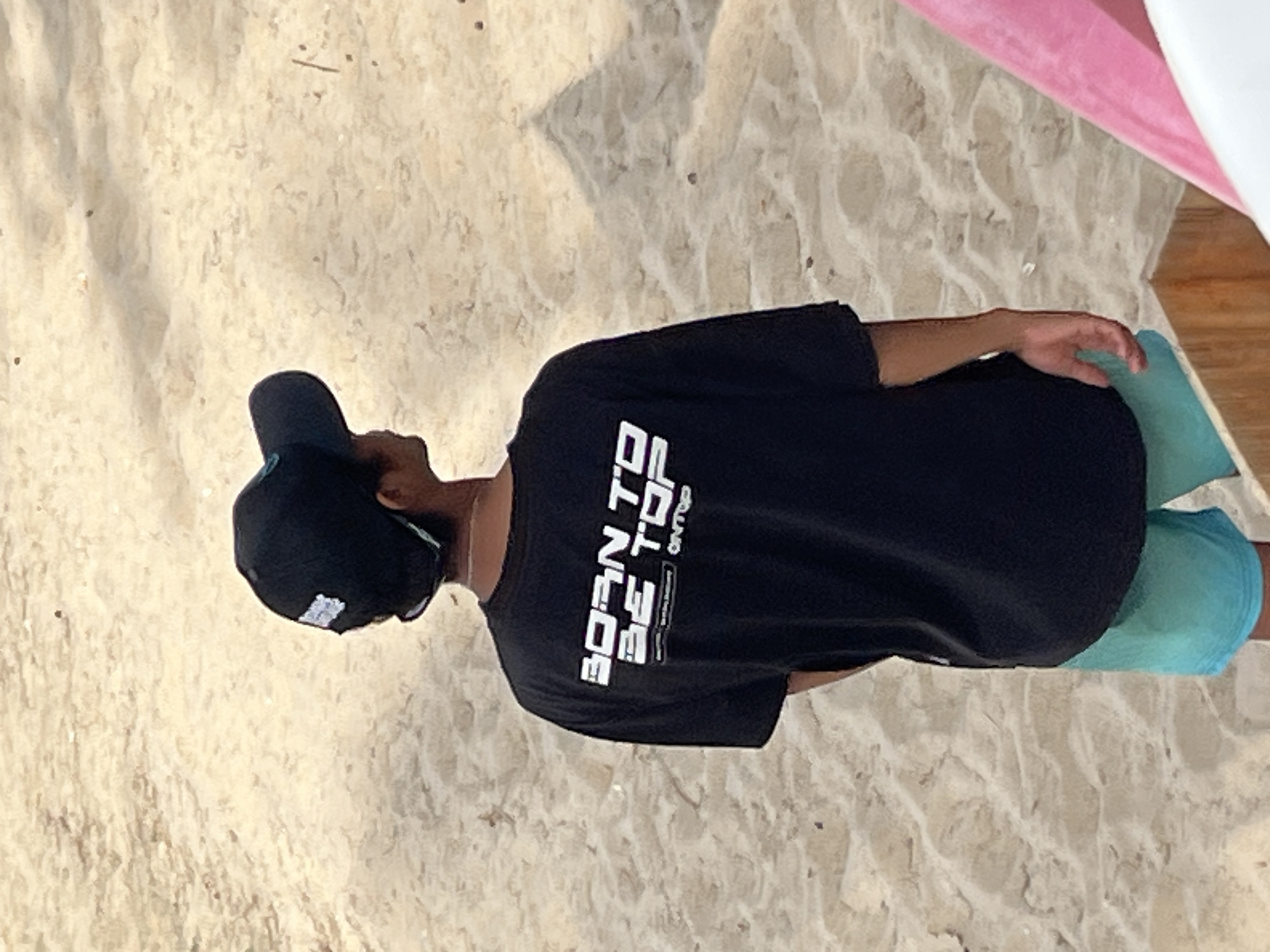 A person wearing a black t-shirt and blue shorts stands on a sandy beach in Phu Thuy Ward, Vietnam. The t-shirt has white text that reads "BORN TO BE TOP" and "ON TOP". The person is also wearing a black baseball cap. The background is a sandy beach with some small objects scattered on it. A portion of a pink object and a wooden structure are visible in the lower right corner of the image. The lighting suggests it is daytime.