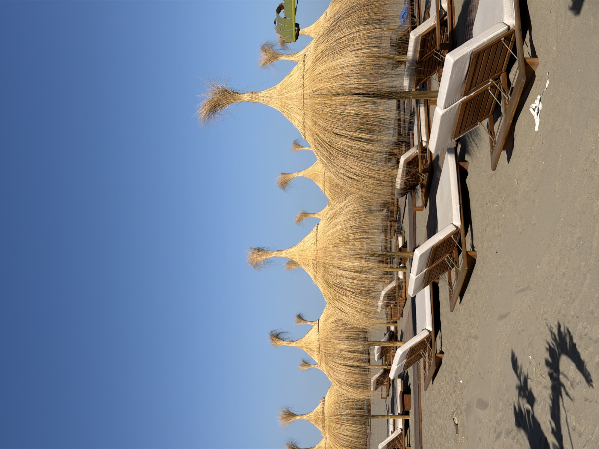 This is a photograph of a beach resort in Playa Del Sol-Villacana, Spain, under a clear blue sky. The scene features rows of empty, straw-thatched umbrellas and lounge chairs set up on a sandy beach. The umbrellas are tall and cone-shaped with a tuft of straw at the top. The lounge chairs are made of wood with white cushions. The beach appears to be unoccupied, with no people visible. The lighting suggests it is daytime, with some shadows cast on the sand. The overall impression is one of tranquility and preparation for visitors.