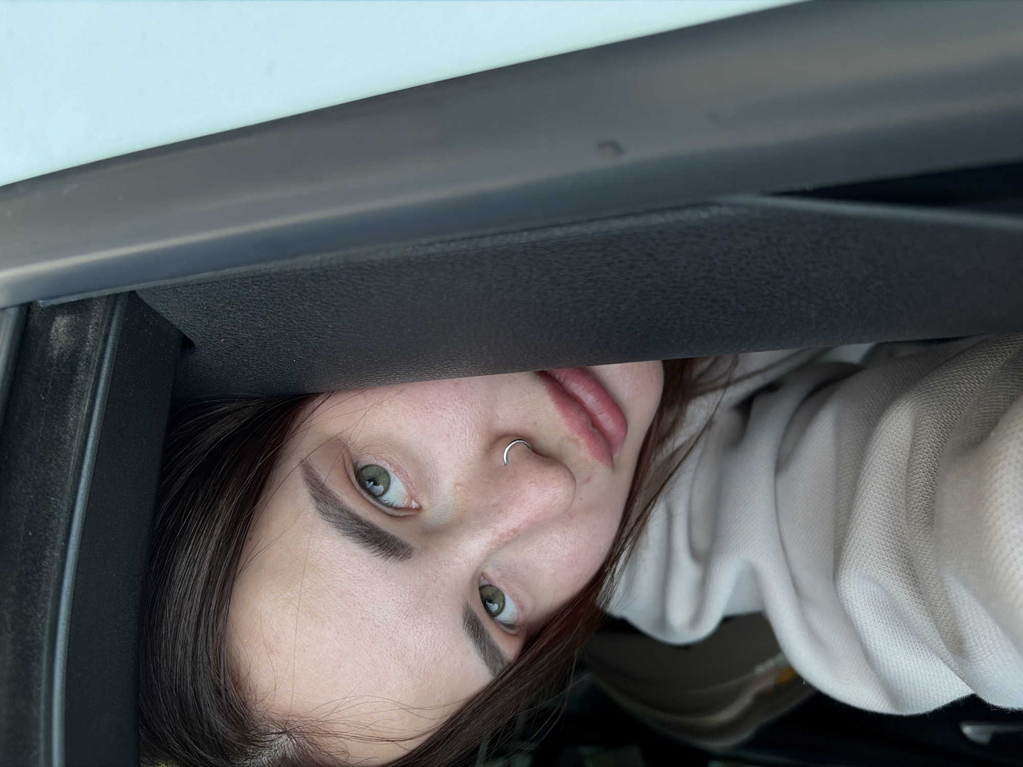 A young woman with fair skin, green eyes, and a nose ring is visible. She is wearing a light-colored hoodie. She appears to be partially obscured by a dark, possibly automotive, dashboard or interior trim. The photo's angle is unusual, suggesting it was taken from a low vantage point, potentially inside a car. There is no information available from the image to confirm the location as Krasnodar, Russia.