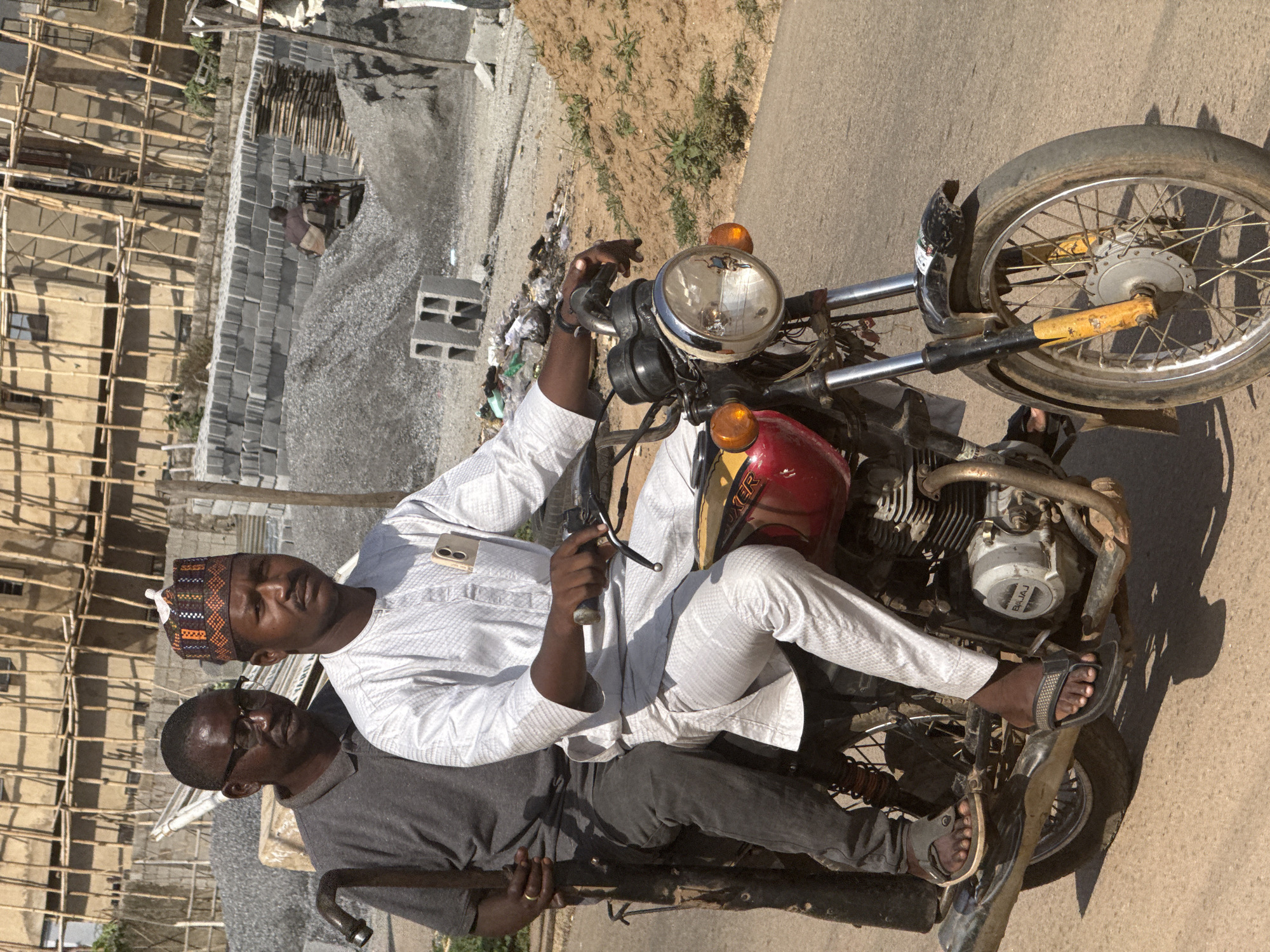 This portrait-oriented image, set in Abuja, Nigeria, captures two men and a motorcycle in a construction or industrial area under bright daylight.
The primary subject is a man seated on a red Bajaj Boxer motorcycle, looking directly at the viewer. He wears a white, textured long-sleeved traditional top, white trousers, and a patterned cap featuring brown, orange, and white geometric designs. A white smartphone, with a visible Samsung logo on its case, is tucked into his shirt pocket. His bare feet, clad in dark open-toed sandals, rest near the motorcycle's footpegs. He holds the handlebar with his right hand.
Partially visible behind and to the left of the seated man is a second man, wearing a dark grey short-sleeved shirt and glasses. He holds a curved metal bar and also looks towards the camera with a neutral expression. He is also wearing dark sandals.
The motorcycle features a chrome headlight, orange turn signals, and a dark red fuel tank with "BOXER" branding. The engine visibly displays the "BAJAJ" brand name. The front wheel has chrome spokes.
The background depicts a developing scene. To the left, there's a building under construction, characterized by extensive wooden scaffolding. In the mid-ground, numerous grey concrete blocks are neatly stacked, alongside piles of gravel or rubble. A small piece of machinery is also visible. The ground is dry and dusty, with sparse vegetation, suggesting a dirt road or pathway. The clear, bright sunlight casts defined shadows, indicating it is either morning or afternoon. The overall setting points to an outdoor, possibly industrial or residential development area.