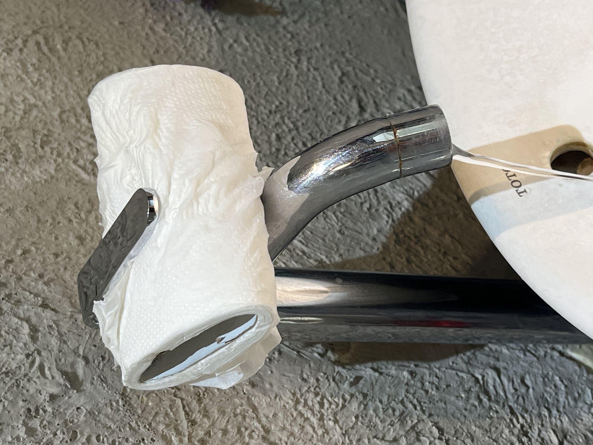 A partially used roll of white toilet paper hangs on a chrome toilet paper holder attached to a white toilet. The scene is located in a bathroom with a textured, gray wall. The toilet is labeled "TOT." The setting is likely Kuta Selatan, Indonesia, based on the location request.