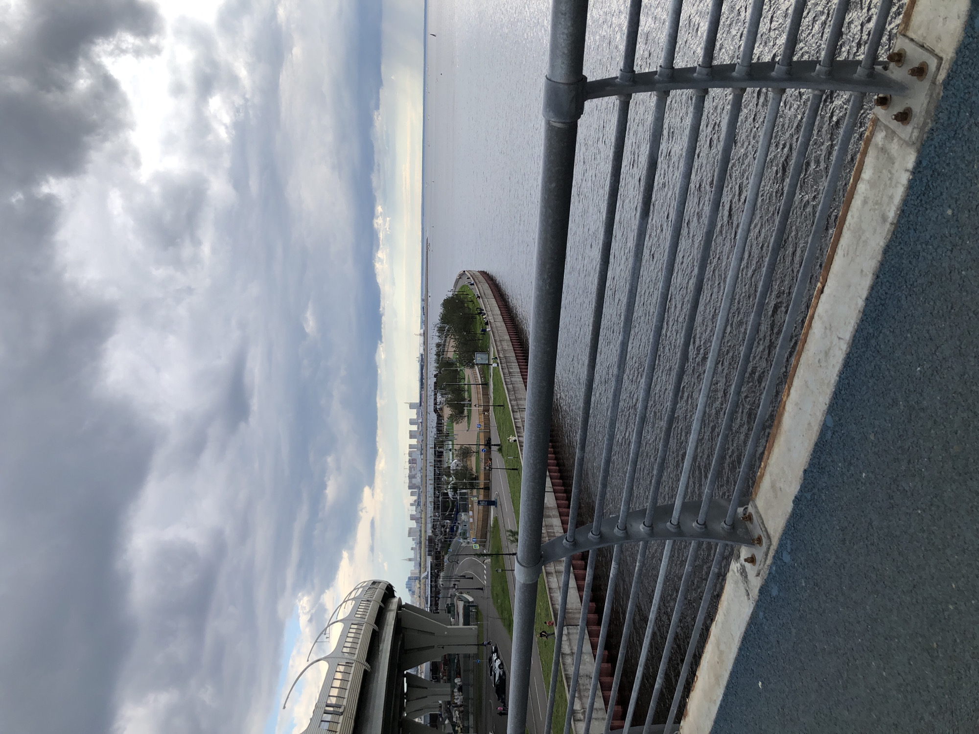 An elevated perspective captures a cloudy, overcast day over a body of water with a cityscape in the distance. In the foreground, a metal railing with horizontal bars is prominent, with a blue paved surface below it. To the left, a curved overpass structure, supported by large concrete pillars, dominates the scene. A road runs beneath the overpass, with parked cars visible. Beyond the road, a landscaped embankment with trees and a pedestrian walkway lines the water's edge. In the middle ground, a row of modern buildings forms a distant skyline. The water itself is calm, with a few small boats visible on the horizon. The overall impression is one of urban infrastructure meeting a natural waterfront under a dramatic sky.