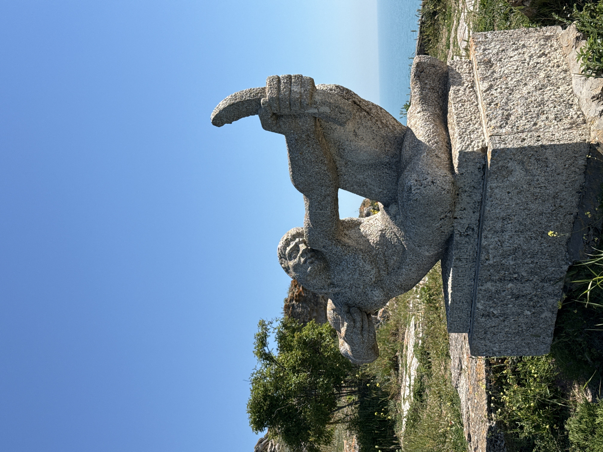 The image captures a stone statue located in Balgarevo, Bulgaria. The statue depicts a male figure lying on his side with one arm raised and holding what appears to be a curved object. The statue rests on a stone pedestal, which is situated on a grassy area. The background features the blue sky and a glimpse of the sea. A tree is visible near the statue. The environment appears to be a coastal area with natural vegetation.