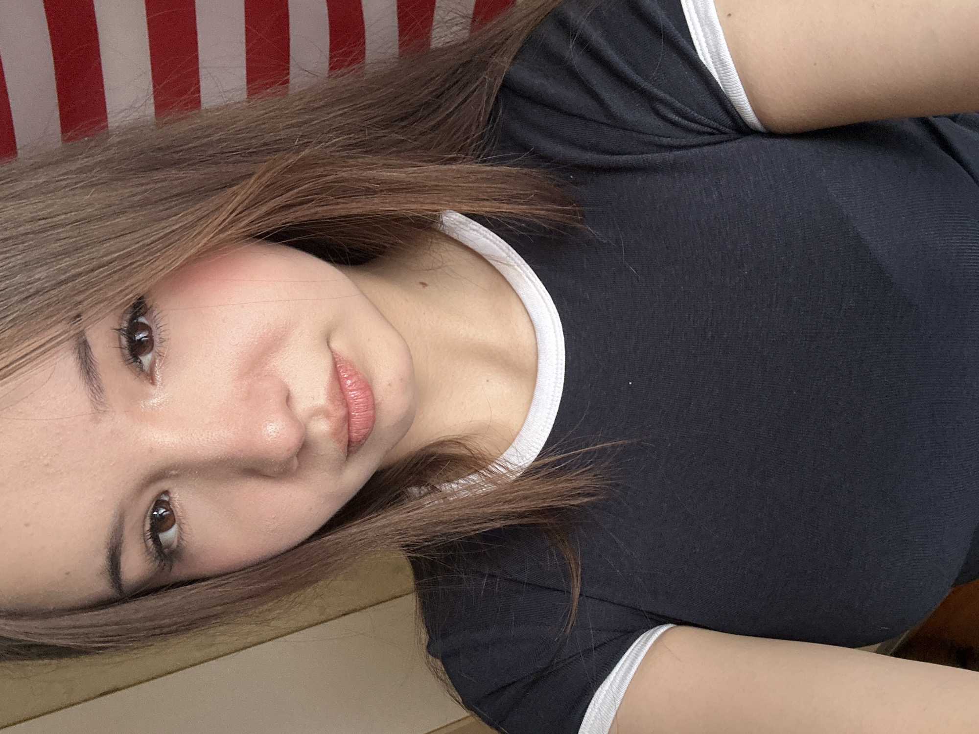This is a close-up selfie of a young woman with long, brown hair. She is wearing a dark t-shirt with white trim on the collar and sleeves. Her skin is fair, and she is wearing light makeup, including mascara and lip gloss. She has brown eyes and her expression is neutral. In the background, to her right, is a red and white striped object, possibly a flag or a banner. To her left, there is a muted, light-colored surface. The lighting appears to be natural, coming from the front. The image is taken indoors, and there are no discernible landmarks or features that would indicate a specific location in Naberezhnye Chelny, Russia.