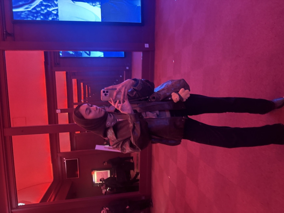 A person wearing a hijab and a jacket takes a selfie in a mirrored space illuminated with red and blue lights. They are holding a phone in their right hand, positioned in front of their face. A purse hangs from their shoulder. Dark pants complete the outfit. The background shows an installation with screens, walls, and reflections creating an endless perspective. Several other figures are visible in the mirror.
