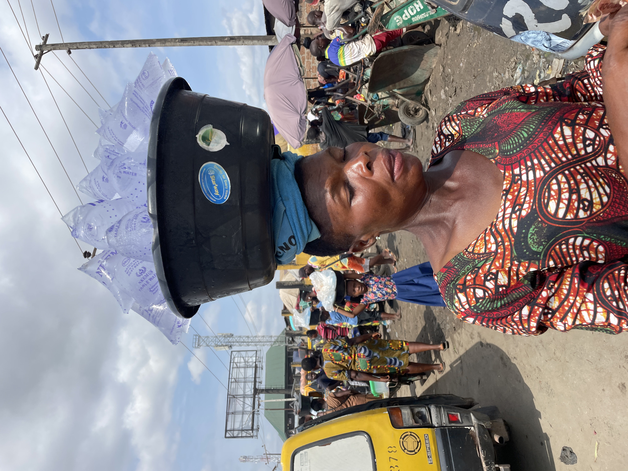 A woman carries a large black tub filled with sachets of water on her head in a busy marketplace. She is wearing a colorful, patterned dress with red, green, and black designs. Her eyes are closed, and her head is tilted back, suggesting exertion or weariness. A blue towel is wrapped around her neck, partially visible under the tub.
The scene appears to be an outdoor market in Fegge, Nigeria, with a dusty ground and various stalls and people in the background. There are other individuals, including a child with a basket on her head, and some carrying goods. A yellow motorized rickshaw is parked in the foreground. Utility poles and wires are visible overhead, and the sky is partly cloudy, indicating it is daytime. Some text is visible on the water sachets, including "WATER", "Starway", and "NKEMUS TANLE WAT". There is also text on the rickshaw, reading "378" and "5-SPEED". The overall impression is one of daily life and commerce in a vibrant, possibly hot, environment.