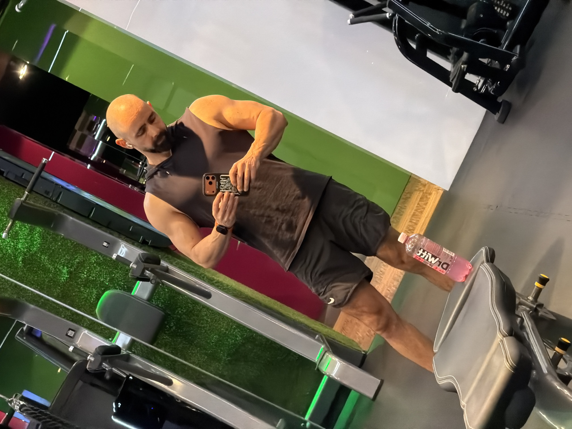 A bald, muscular man with a beard is taking a selfie in what appears to be a gym. He is wearing a dark gray sleeveless shirt, black shorts, and a watch. The gym has green and white walls, and there is gym equipment in the background, including a rowing machine and a weight machine. A water bottle is on a seat. The location in the city of Plovdiv, Bulgaria cannot be verified.