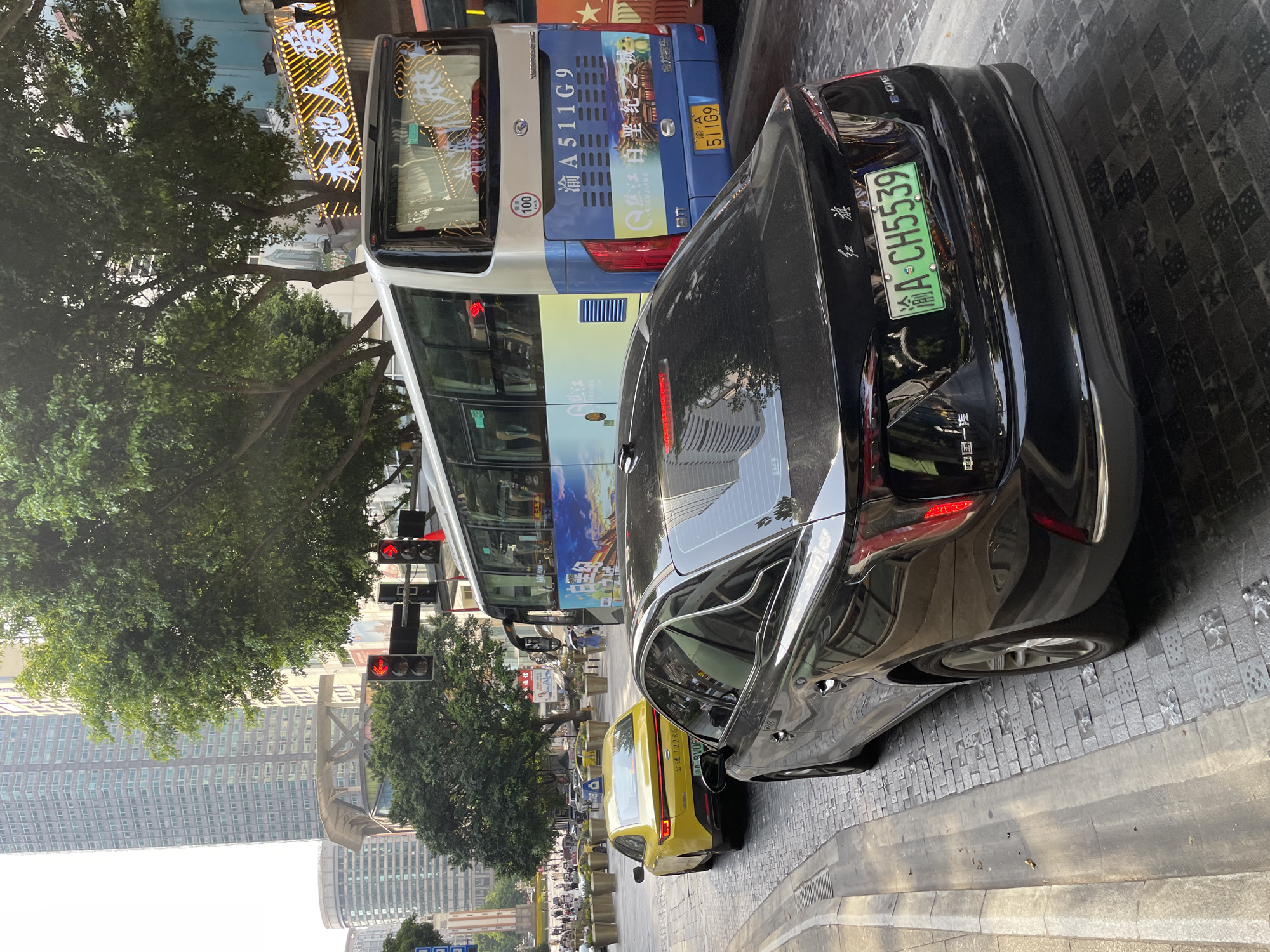 The image captures a street scene in Yuzhong District, China, during the daytime. A black sedan is parked on the right side of a cobblestone road, facing away from the camera. The sedan has a green license plate that reads "渝A CH5539". To the left of the black sedan, a yellow sedan is also parked. A large blue and white bus is in motion, positioned behind the yellow sedan and to the right of the black sedan, suggesting traffic flow. The bus has a license plate that reads "渝A 511G9". The background reveals tall buildings, lush green trees, and traffic lights with red signals illuminated. Pedestrians are visible on a sidewalk in the distance. The overall environment appears to be an urban setting with commercial and residential elements.