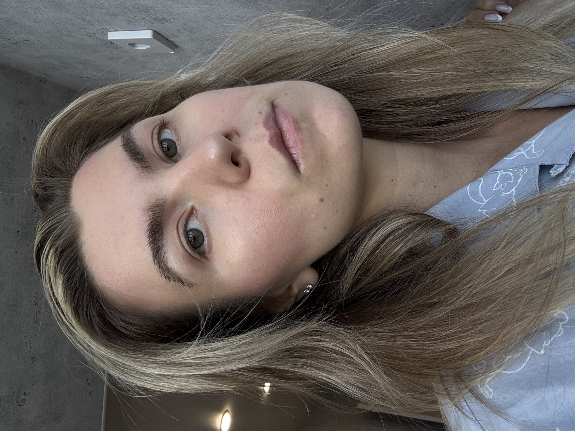 A close-up, angled portrait shows a woman with light blonde hair and green eyes. She is wearing a light blue patterned top. The background consists of a gray wall with a white electrical fixture near the top. The woman is facing the camera, with her head tilted to the side. She appears to be indoors under artificial lighting. There is no indication of a city or specific location.