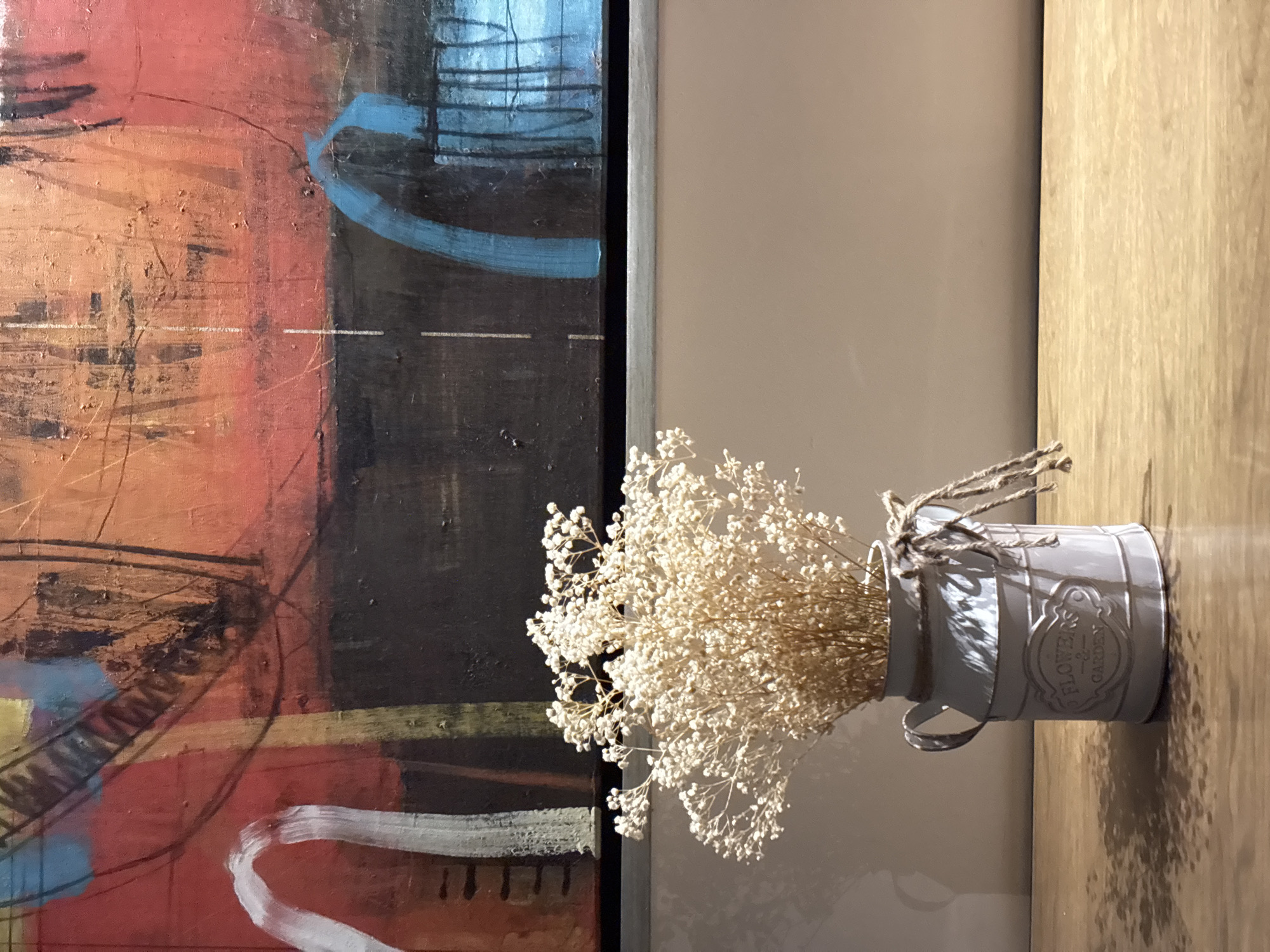 The image shows a still life arrangement. On the left, an abstract painting hangs, featuring a mix of colors including red, orange, blue, and black. Next to the painting, a light grey, galvanized metal container sits on a wooden surface. The container is labeled "FLOWERS GARDEN" and holds a bouquet of dried baby's breath flowers. The background consists of a light beige wall. The location, San Bernardino Tlaxcalancingo, Mexico, is not directly evidenced in the photograph and is only assumed based on the prompt. No people or actions are visible.