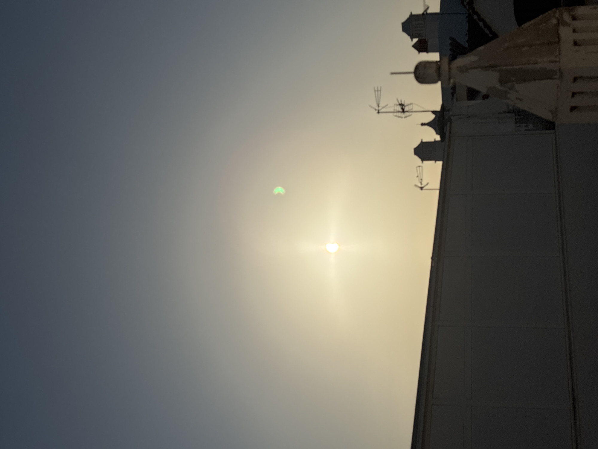 The image captures a clear sky above a rooftop scene in Tavira, Portugal. The sun, partially eclipsed, is visible as a bright orb with a halo of light, positioned slightly to the left of center in the upper half of the frame. To the left of the sun, a crescent-shaped greenish light artifact is present, likely a lens flare or atmospheric phenomenon. Silhouetted against the sky are several rooftop structures, including what appear to be chimneys and a distinct, ornate spire in the foreground on the right. In the mid-ground, a long, flat-topped structure runs horizontally across the lower portion of the image. The overall lighting suggests either sunrise or sunset, with a gradient of light tones in the sky. There are no people or animals visible in the image.