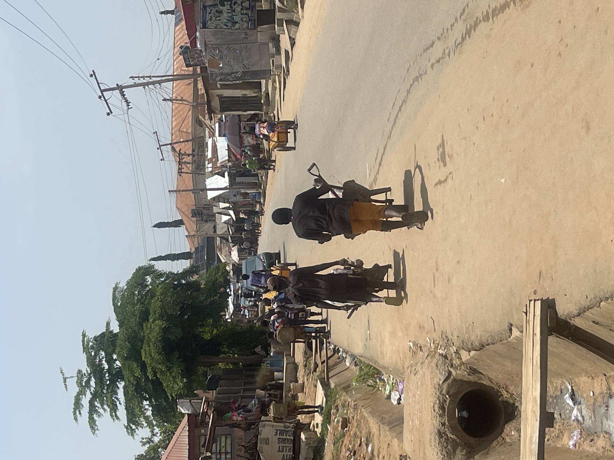 The image depicts a street scene in Abuja, Nigeria, likely in a bustling commercial or residential area. The weather appears to be sunny and clear, contributing to the bright lighting of the scene.
In the foreground, two young men are walking along a paved road, pushing a wheelbarrow between them. They are both dressed casually, with one wearing a black t-shirt and orange shorts, and the other in a dark t-shirt and shorts. The wheelbarrow appears to be loaded with some items, though their exact nature is not discernible. They are walking away from the viewer, towards the center of the image.
To the left of the road, there is a drainage ditch that runs alongside the sidewalk. The ditch is partially filled with water and debris, indicating potential sanitation challenges. A makeshift wooden plank bridges a section of the ditch. Adjacent to the ditch are various structures, including shops and residential buildings with corrugated iron roofs. Some of these buildings display signage, though the text is mostly illegible.
Further down the street, other individuals and vehicles are visible. A few people are walking or cycling, and a dark-colored car is seen in the distance. There are also several utility poles with a complex network of wires crisscrossing the sky, a common sight in urban areas. The buildings lining the street are a mix of single-story and multi-story structures, some appearing to be shops or market stalls.
The overall atmosphere of the image suggests a typical day in a vibrant urban environment, with people engaged in their daily activities, transporting goods, and navigating the local infrastructure. The presence of the drainage ditch and the visible debris hints at some of the environmental conditions in the area.
