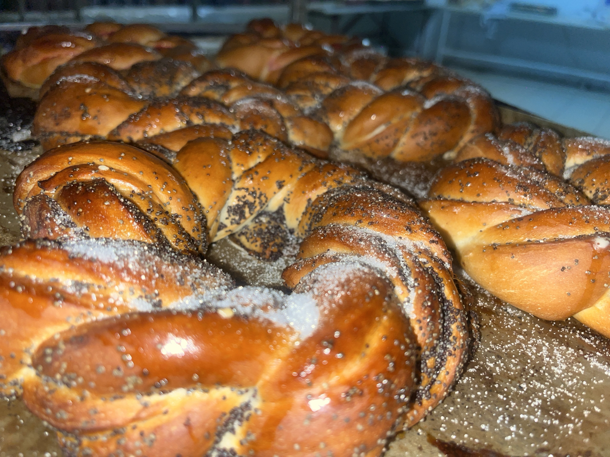 A close-up shot depicts several braided loaves of bread on a baking sheet. The loaves are golden brown, sprinkled with poppy seeds and coarse sugar. They appear to be fresh out of the oven. The location in Novopokrovskaya, Russia is not discernible from the image.