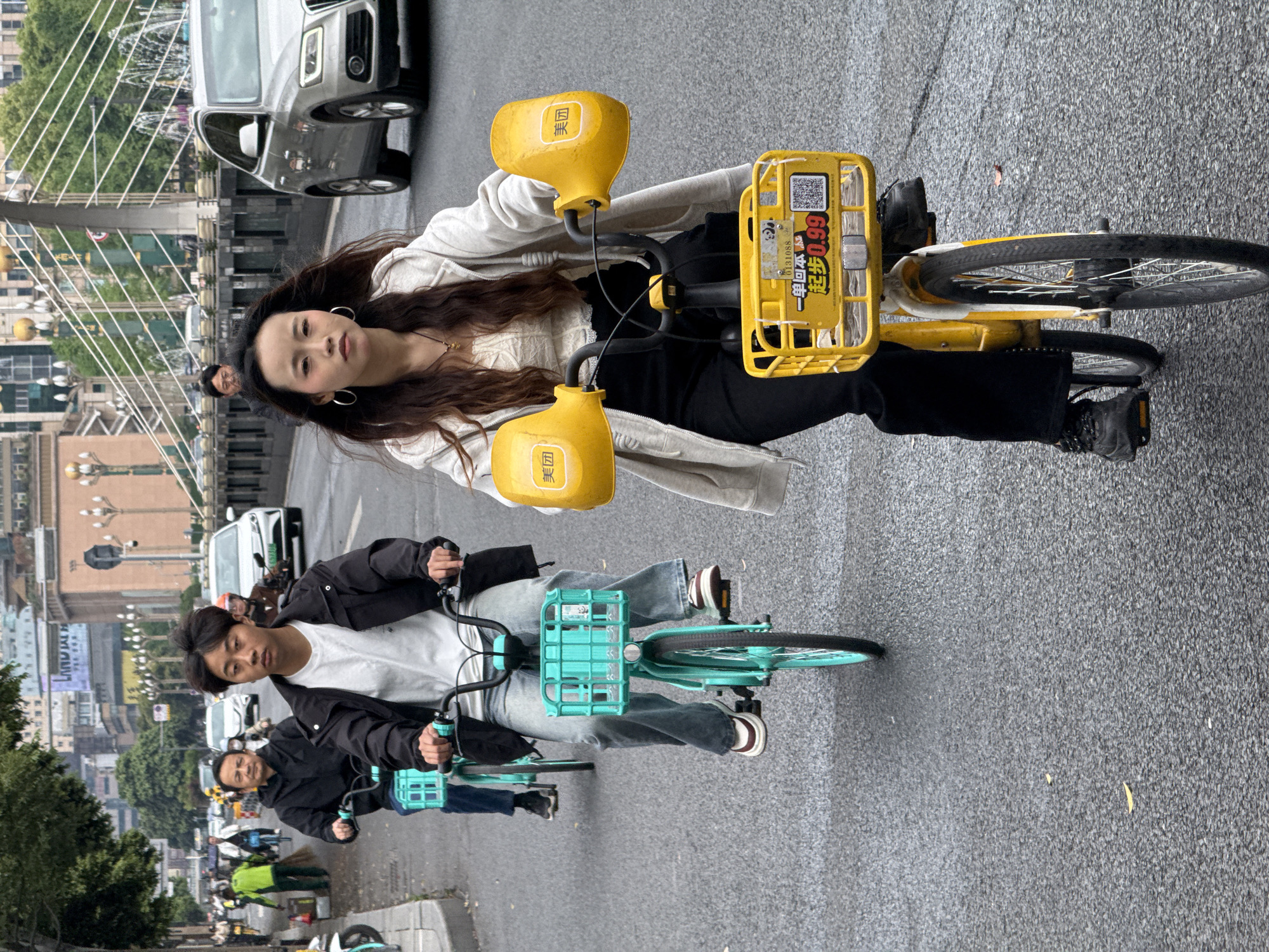 The image shows a street scene in Qingyang District, China. A young woman with long brown hair is riding a yellow shared bicycle towards the camera. She is wearing a white sweater, black pants, and silver hoop earrings. Behind her, a man is riding a turquoise shared bicycle. He is wearing a black jacket, a white T-shirt, and jeans. A few more cyclists can be seen behind him, receding into the distance. The street is wet and gray, suggesting recent rain. Buildings, trees, and a bridge are visible in the background. Cars are also visible on the bridge.