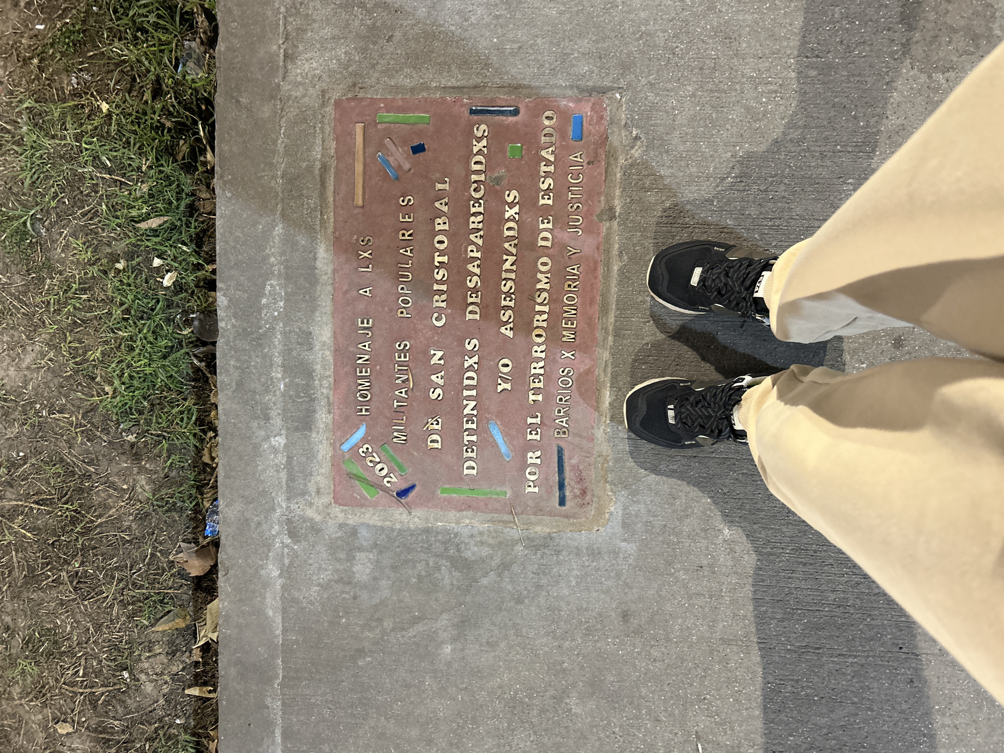 The image shows a memorial plaque set into a concrete sidewalk in Buenos Aires, Argentina. The plaque is rectangular and made of a reddish material with inlaid tiles. The inscription on the plaque reads: "HOMENAJE A LXS MILITANTES POPULARES DE SAN CRISTOBAL DETENIDXS DESAPARECIDXS Y/O ASESINADXS POR EL TERRORISMO DE ESTADO BARRIOS X MEMORIA Y JUSTICIA". The year "2023" is also visible. In the foreground, a person's feet wearing black sneakers and light-colored pants are visible, indicating the perspective is from someone standing and looking down at the memorial. The ground around the memorial appears to be a sidewalk and a grassy area with some fallen leaves.