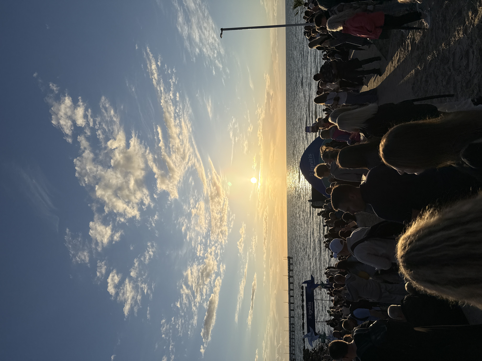 A crowd of people are gathered on a beach at sunset. The sun is low on the horizon, casting a warm glow across the scene. The ocean is calm, with gentle waves lapping at the shore. In the distance, a pier stretches out into the water. There are several tents and structures set up on the beach, suggesting an event or festival is taking place. The sky is a mix of blue and orange, with scattered clouds. The people in the crowd are diverse in age and appearance, and they appear to be enjoying the sunset and the atmosphere. Some are standing, while others are sitting on the sand. The overall impression is one of a peaceful and festive gathering by the sea.