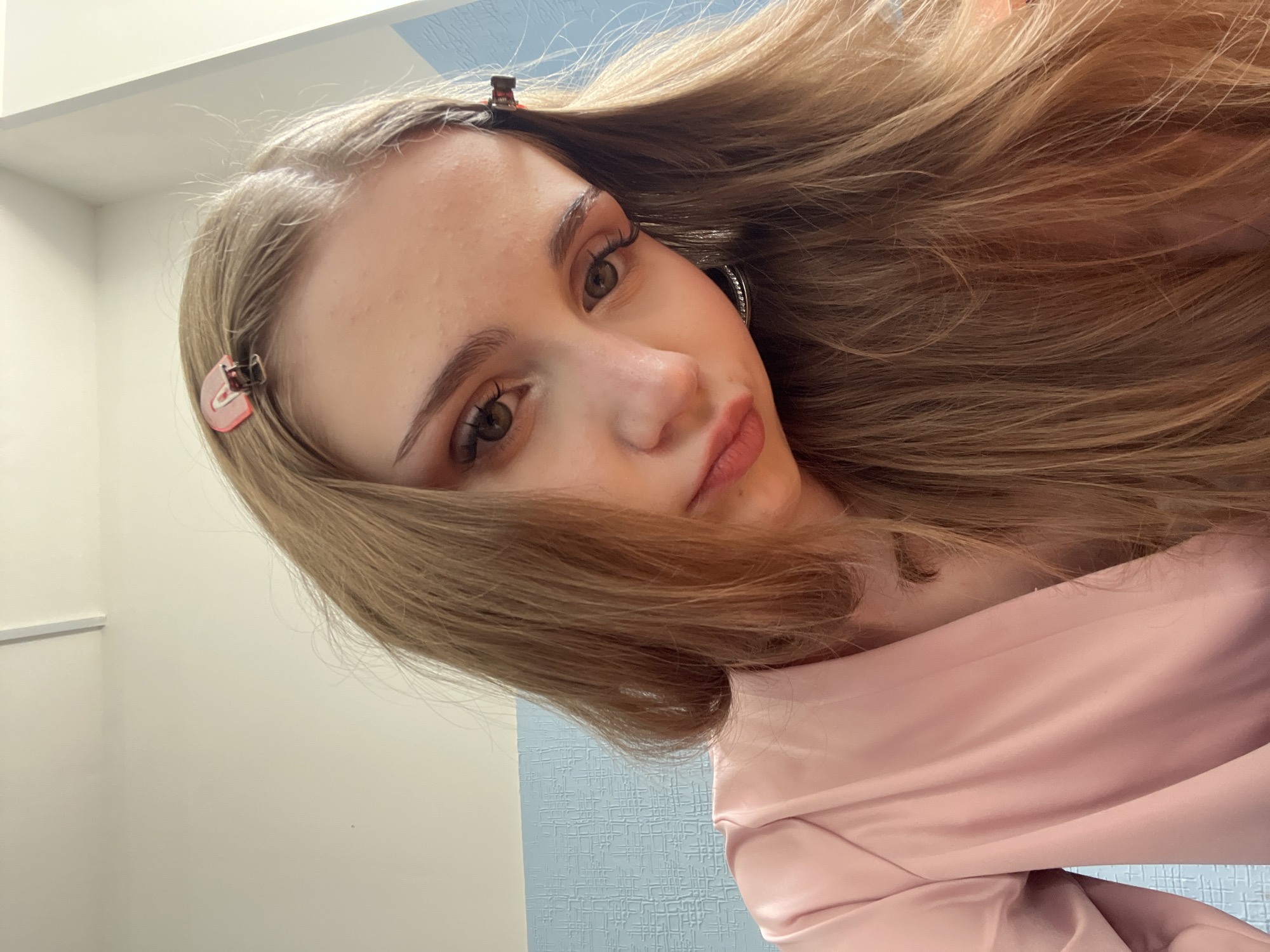 A close-up selfie shows a young woman with light brown hair and light skin, wearing a pink satin robe. She has makeup on, including eyeliner and eyeshadow, and her lips are slightly parted. Two hair clips are visible in her hair, one pink and shaped like a heart, and another brown one. She is wearing a large hoop earring with small crystals. The background is a light blue textured wall and a white ceiling. The image appears to have been taken indoors. The location is not identifiable as Sterlitamak, Russia, nor is there any indication of that city in the image.