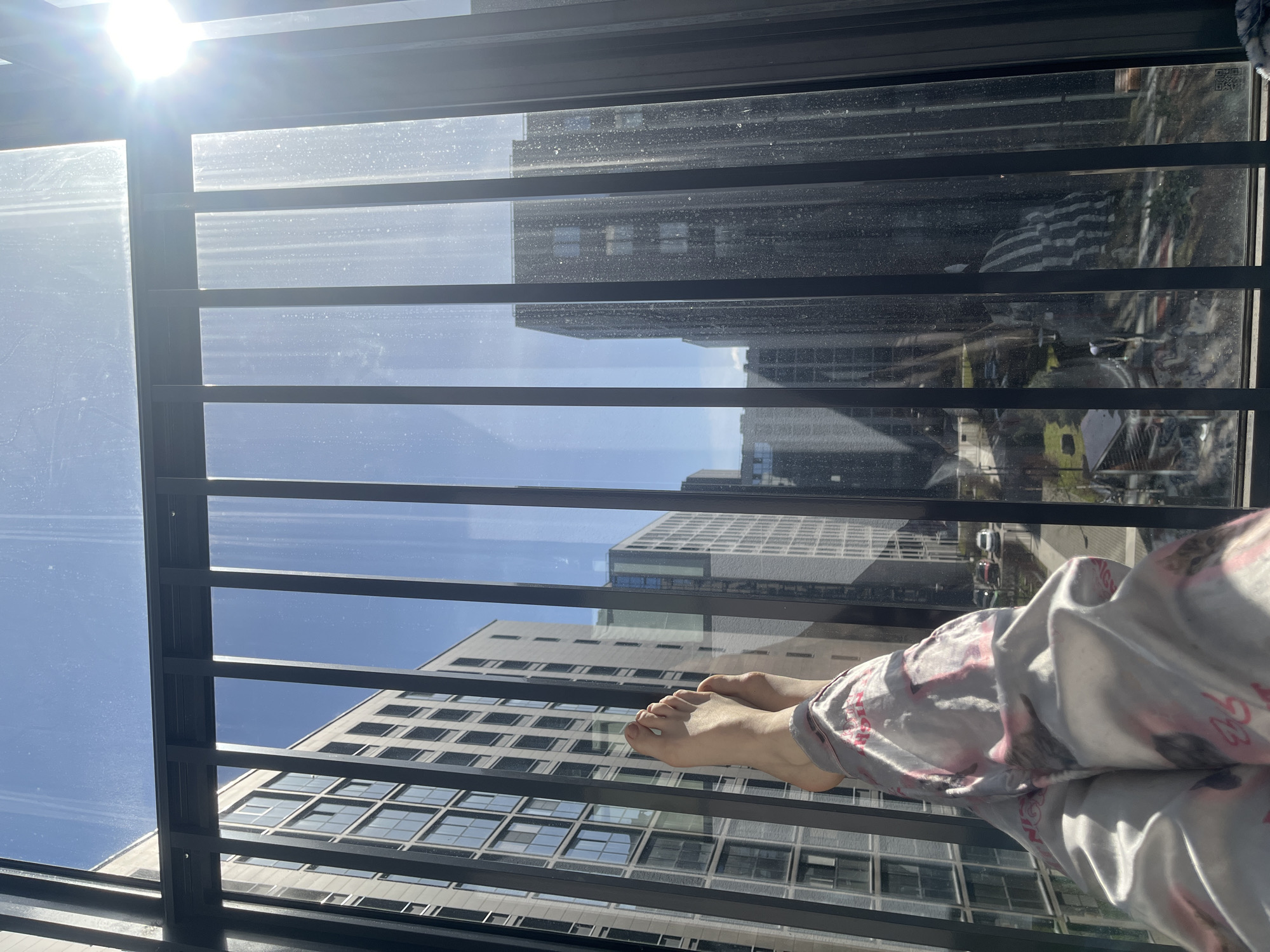 The image depicts a view from a balcony or window in an urban setting, presumably in Saint Petersburg, Russia, based on the building styles visible. The dominant feature is the grid of the balcony railing, which obscures but does not entirely block the view of the buildings across the street. Sunlight glares off the glass panels of the railing, creating bright spots. In the foreground, a person's feet and legs, covered in patterned gray and pink pajamas, are visible. The person appears to be lying down, positioned horizontally in the frame. The buildings in the background consist of various heights and architectural styles. The sky is blue and partially visible through the gaps in the railing and between the buildings.