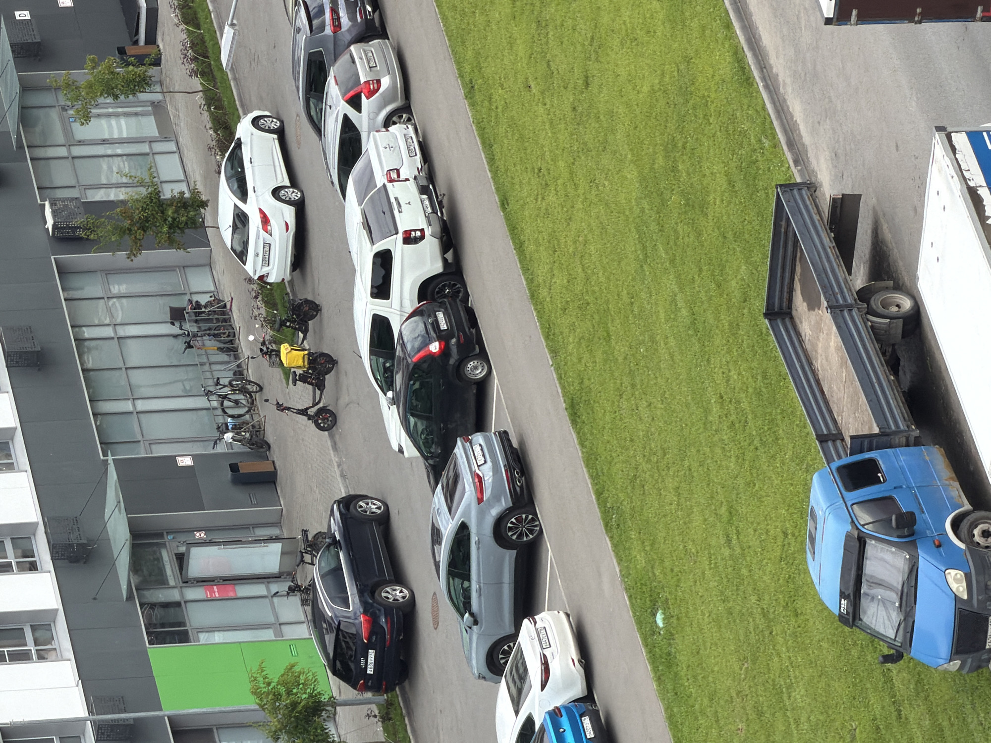 The image shows an outdoor parking lot next to a modern building with large windows. Several vehicles are parked in designated spots, including a dark blue Audi SUV, a light blue sedan, a white sedan, a dark colored sedan, a white pickup truck with a camper shell, a silver hatchback, and a dark grey SUV. Two electric scooters are parked near the building entrance. A blue flatbed truck is parked on the right side of the image. The building appears to be an office or commercial space, with trees and landscaping visible in the background. The ground is paved in the parking lot area and covered with green grass.