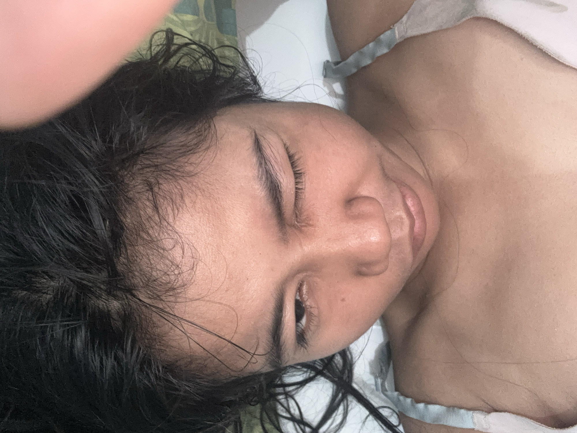 The image is a close-up, selfie-style shot of a young woman's face and upper chest. She has dark, wet hair that is spread around her head. Her eyes are partially closed, with one eye winking. She is wearing a light-colored camisole or bra. The lighting is warm and somewhat dim. The background appears to be indoors, possibly a bedroom, with a patterned fabric visible behind her. There is a blurred pink object, likely a finger, in the upper right corner of the frame. The image has been taken from a high angle looking down at the subject. This image was taken in North Jakarta, Indonesia.
