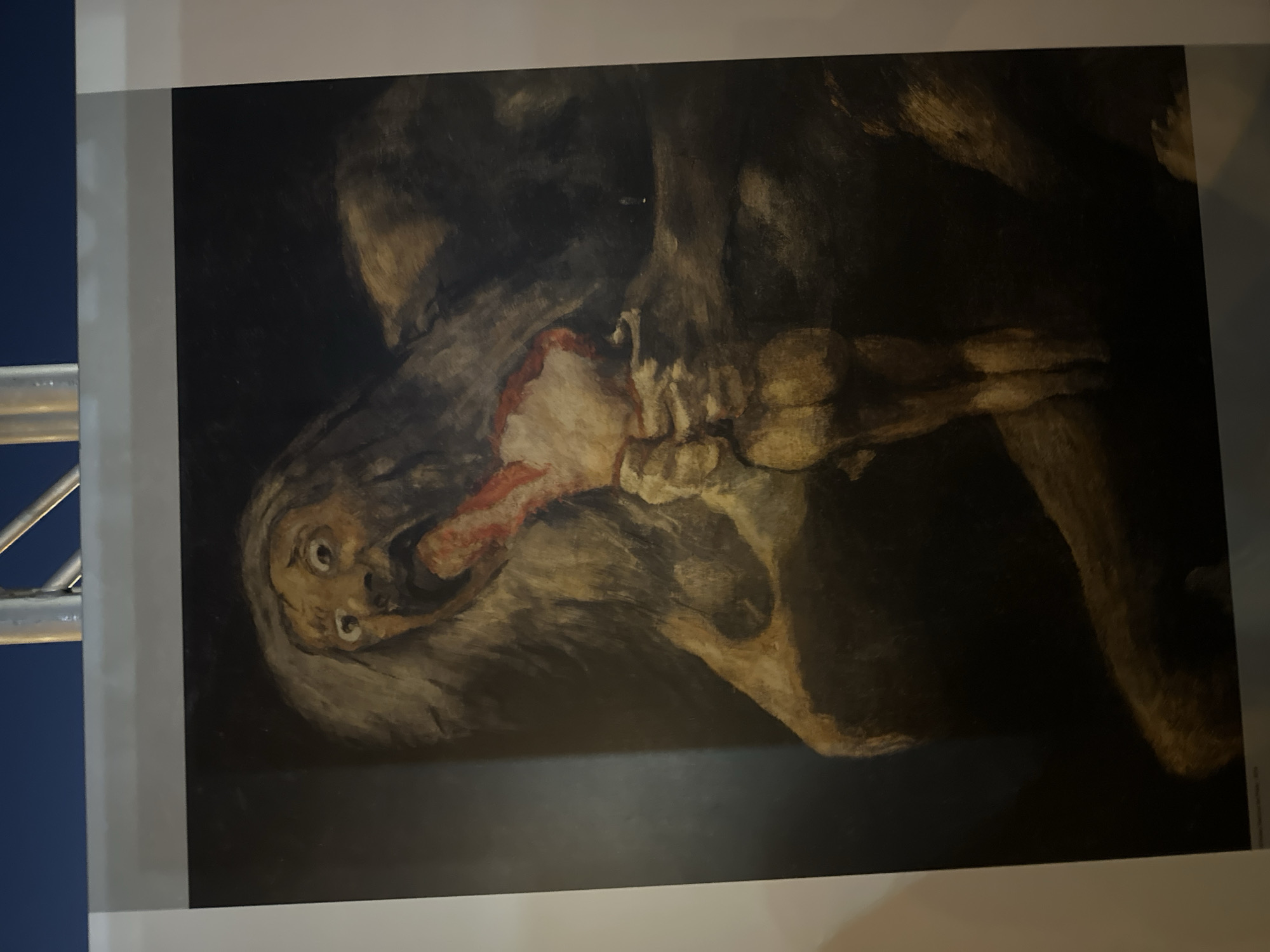 The image shows a painting depicting a monstrous, emaciated figure with long, unkempt hair, hunched over and consuming a severed limb. The figure's mouth is agape, revealing a grotesque tongue and teeth, and its eyes are wide with a manic expression. The scene is dark and atmospheric, with the figures rendered in earthy tones and heavy shadow, suggesting a nightmarish or violent subject matter. The painting appears to be on display, with a portion of a white border and a silver pole visible at the top of the frame, indicating it is likely in a gallery or exhibition. The text at the bottom of the image is partially visible and includes the words "Mazarrón, Spain." This suggests the painting is either located in Mazarrón, Spain, or is an artwork related to the city.