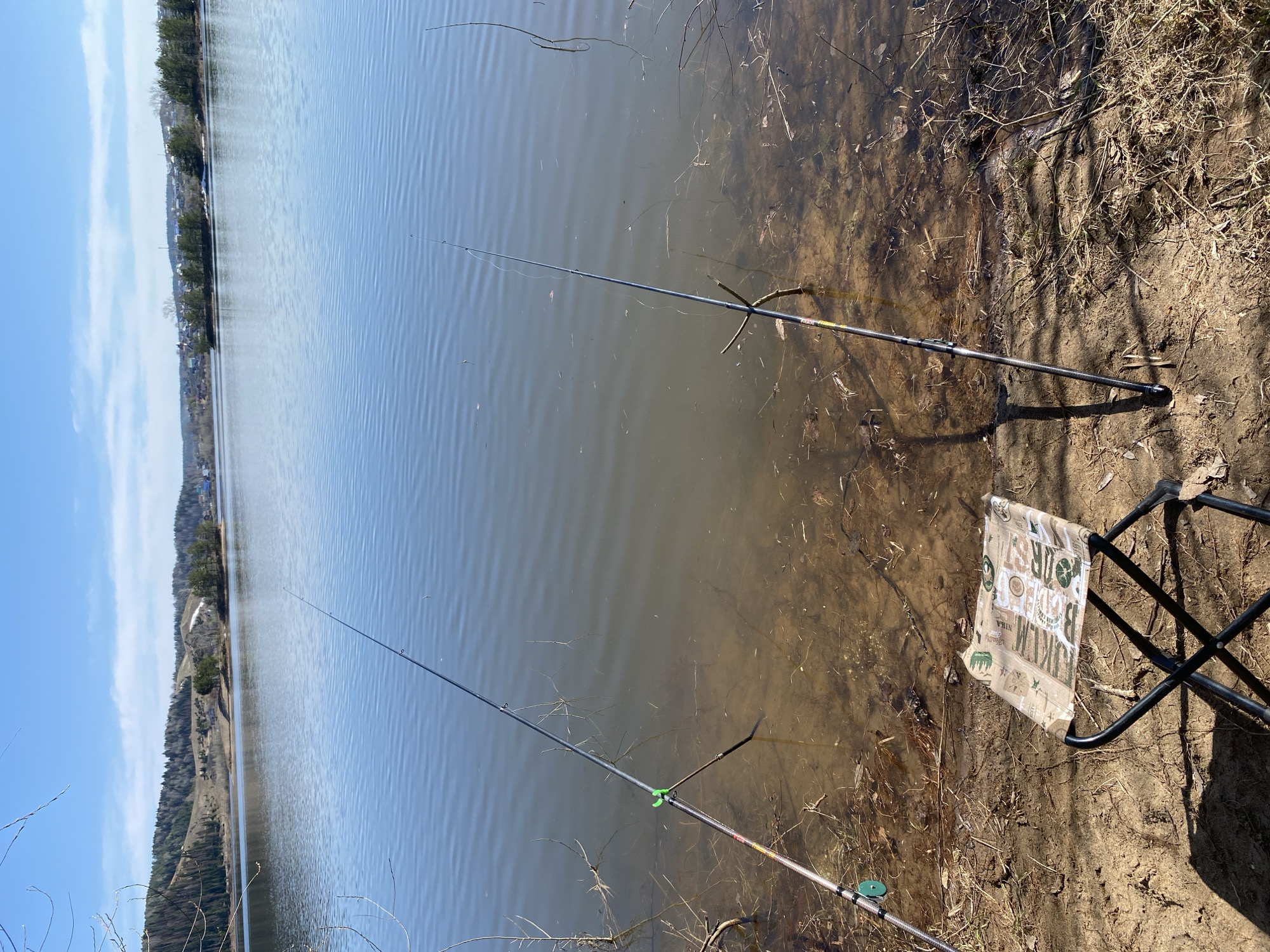 The image shows a lakeside view with two fishing rods set up on the shore. A folding chair sits nearby on the muddy bank. The rods extend into the water, which appears calm with slight ripples. The shoreline is a mix of mud, grass, and small branches. In the background, a forested area is visible, indicating the location is likely in a rural setting near Ольховка, Russia. The sky is blue with some clouds, suggesting it is a clear day. There are no people visible in the image.