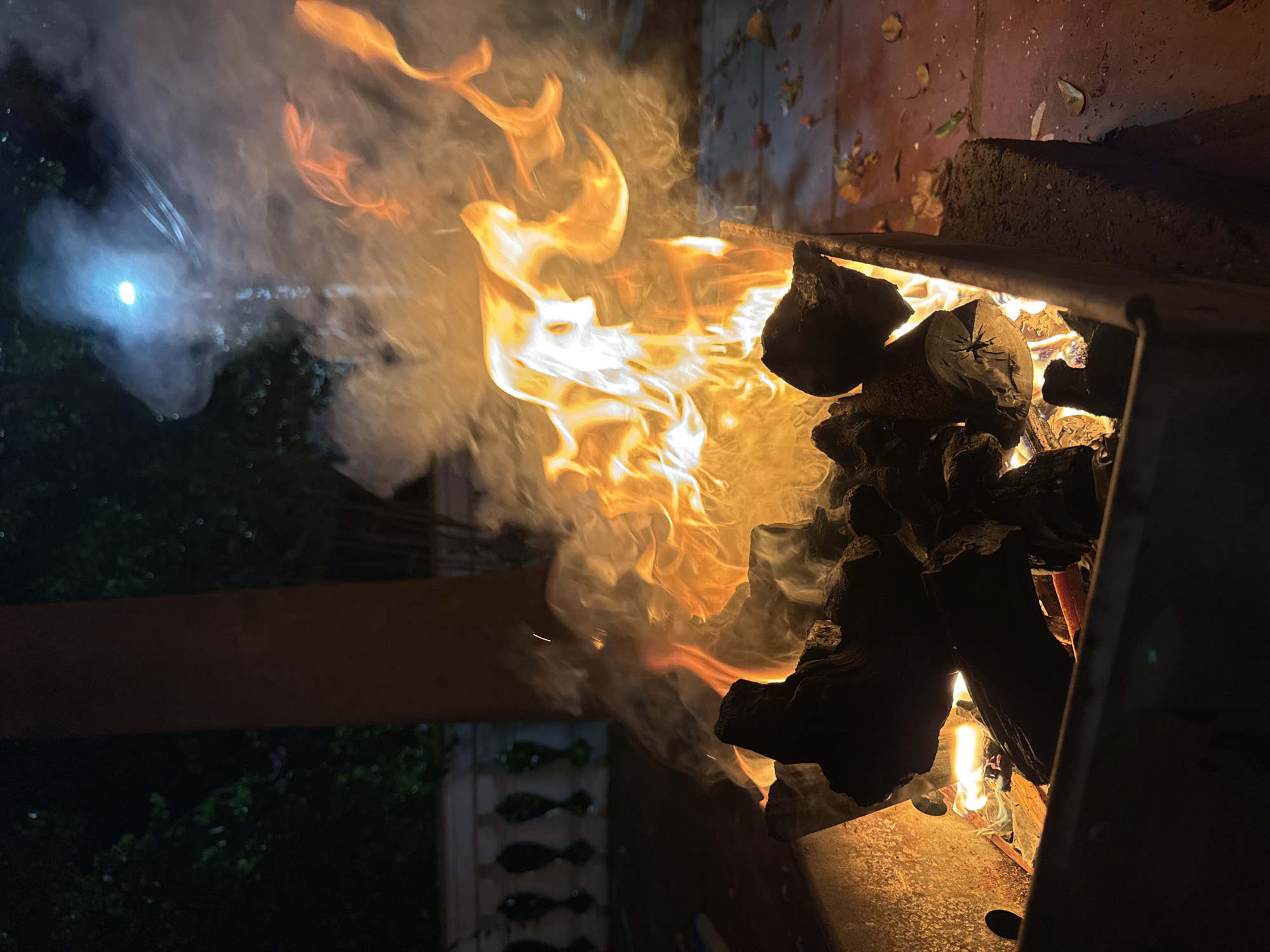 A fire burns in a metal brazier at night, producing a significant amount of smoke and orange flames. The brazier contains charcoal and wood. The background is dark, with trees and foliage visible on the left. A white, ornate railing is present behind the brazier, suggesting an outdoor patio or balcony setting. A bright light source is visible in the upper right portion of the image, illuminating the smoke. The ground around the brazier appears to be paved with red tiles and scattered with fallen leaves. The scene does not depict any individuals or specific locations within Mũi Né, Vietnam.