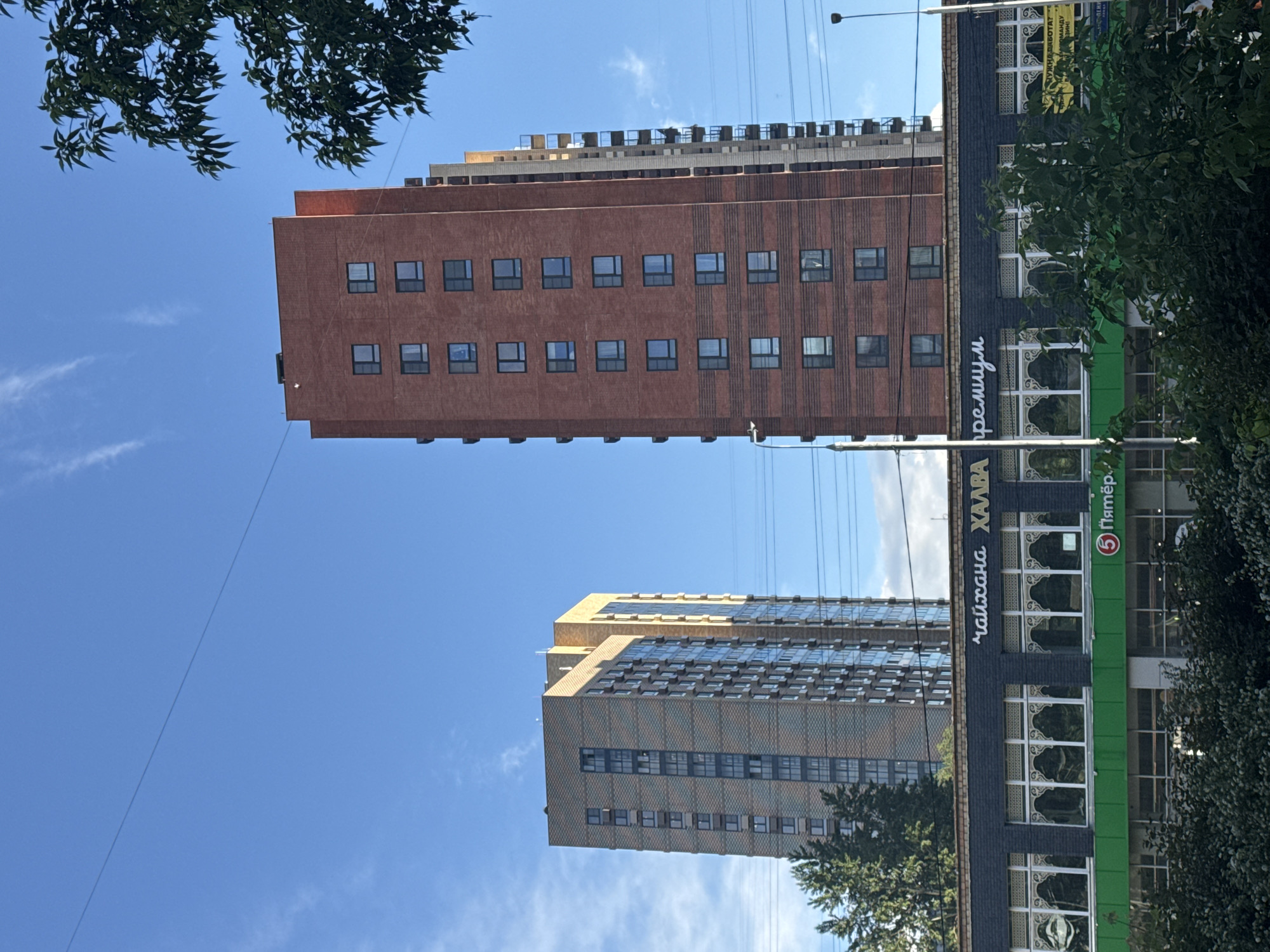 The image shows a view of buildings in Moscow, Russia. Two tall buildings dominate the frame, one primarily brick with multiple rows of windows, and another with a more complex facade featuring reflective windows. In the foreground, a building with a black and green facade displays signs in Russian, including "Чайхана ХАЛВА" (Chaikhana Halva) and "Пятёрочка" (Pyaterochka), a Russian supermarket chain. Green foliage from trees is visible on the sides and in the foreground, partially obscuring some of the buildings. Overhead, electrical wires stretch across the clear blue sky. The buildings appear to be modern apartment or commercial structures.
